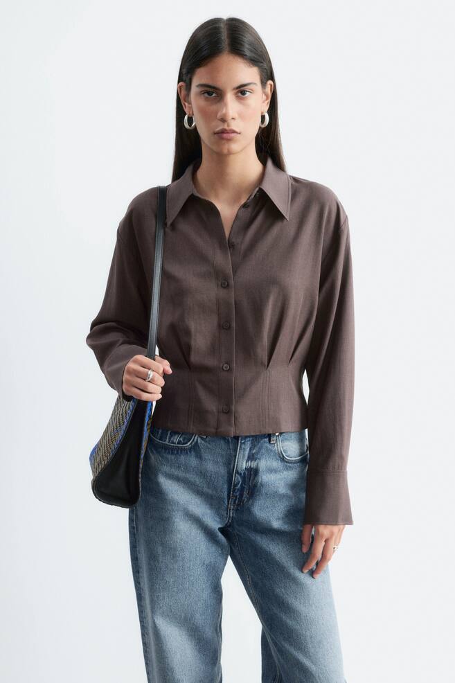 Das Model trÃ¤gt ein braunes Hemd zum ZuknÃ¶pfen mit spitzem Kragen, langen Ãrmeln und Taillenfalten, die die Silhouette formen. Das Hemd ist mit einer blauen Jeans kombiniert, und eine gewebte UmhÃ¤ngetasche wird Ã¼ber der Schulter des Models getragen. - 1