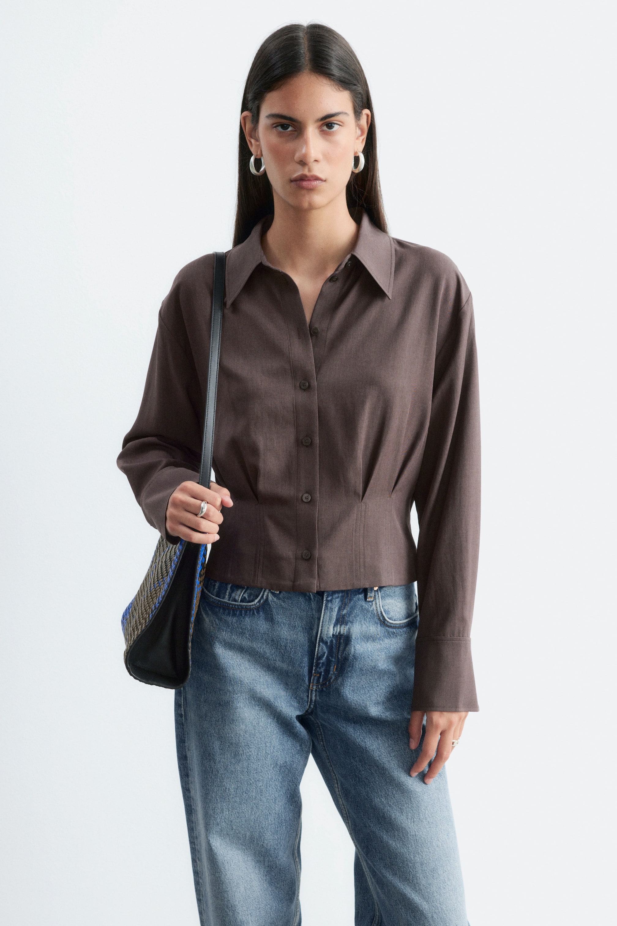 Ver imagem maior: A modelo veste uma camisa castanha com botões, com gola marcada, mangas compridas e pregas na cintura que moldam a silhueta. A camisa é combinada com jeans azuis e uma mala de ombro entrançada é transportada ao ombro da modelo.