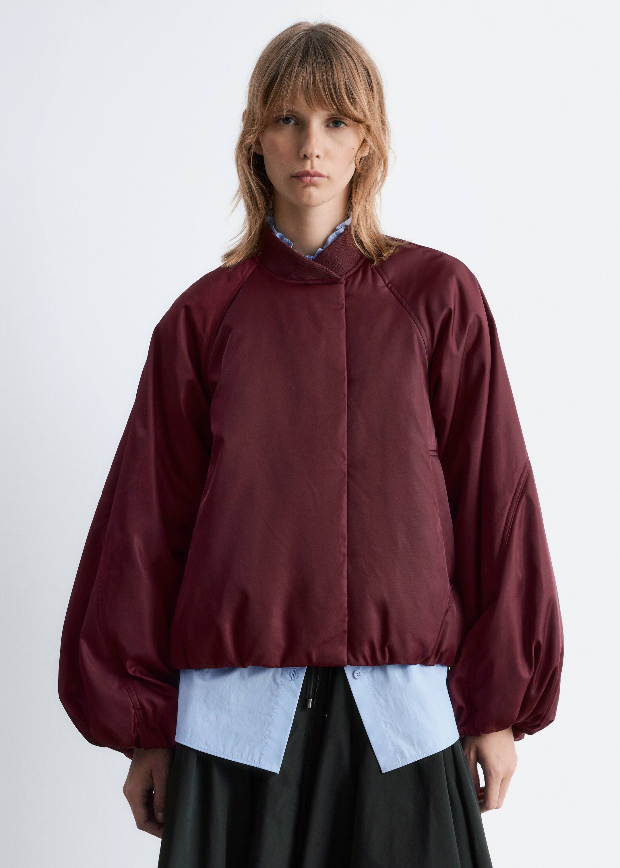 Visualizza immagine più grande: Una giacca bomber bordeaux è in mostra, caratterizzata da un tessuto liscio e leggermente lucido e da una chiusura frontale nascosta. È progettata con un colletto rialzato a coste, maniche raglan voluminose che si stringono ai polsi e un orlo corto a coste. La giacca è sovrapposta a una camicia azzurra con colletto, visibile al collo arricciato e che si estende sotto l'orlo della giacca.