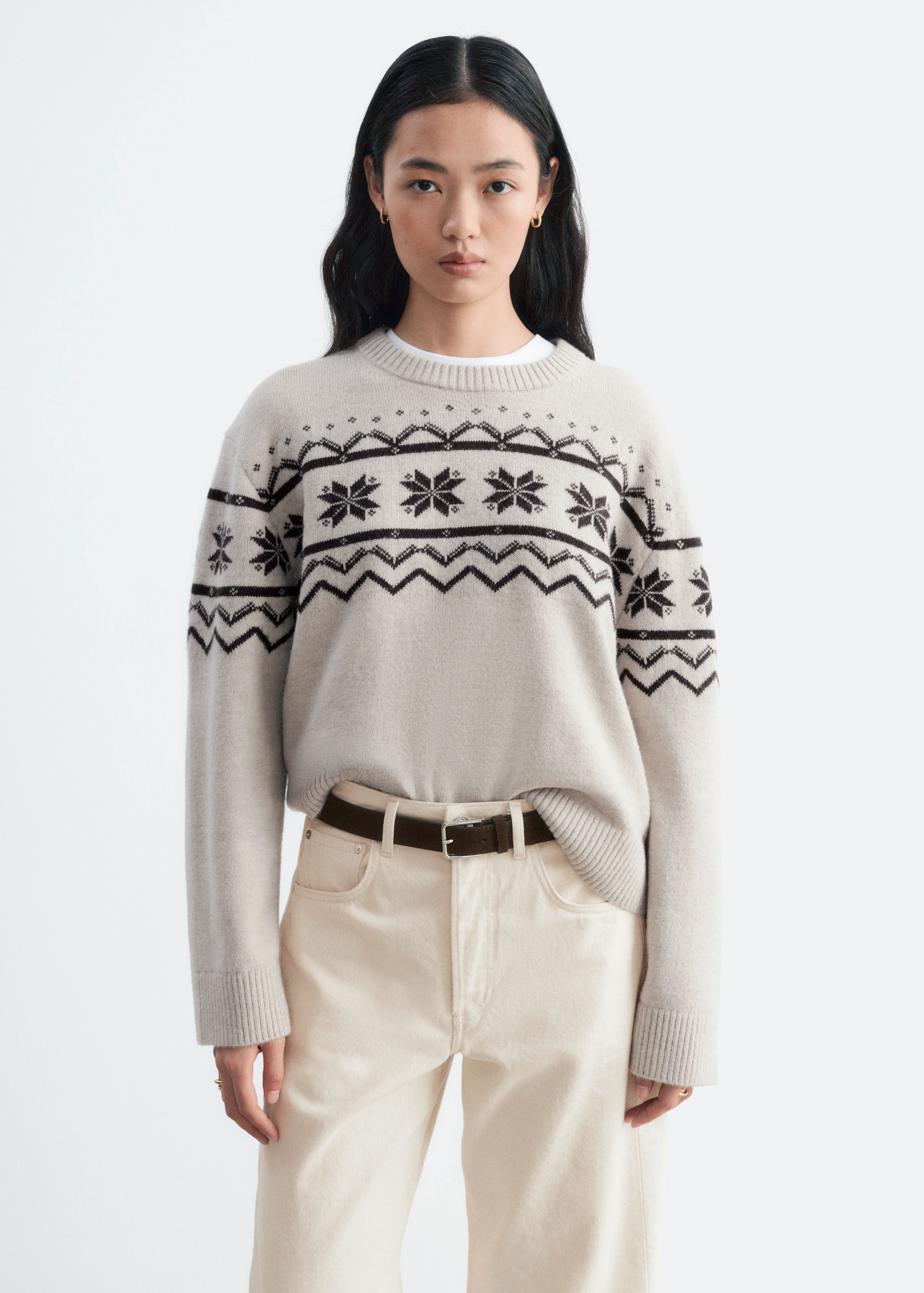JasnobeÅ¼owy sweter dzianinowy jest pokazany od przodu, prezentujÄc ciemnobrÄzowy wzÃ³r Fair Isle ze ÅnieÅ¼ynkami i geometrycznymi motywami na klatce piersiowej oraz gÃ³rnej czÄÅci rÄkawÃ³w. Ubranie ma pudeÅkowy fason wykoÅczony prÄÅ¼kowanym okrÄgÅym dekoltem, prÄÅ¼kowanymi mankietami i prÄÅ¼kowanym doÅem.