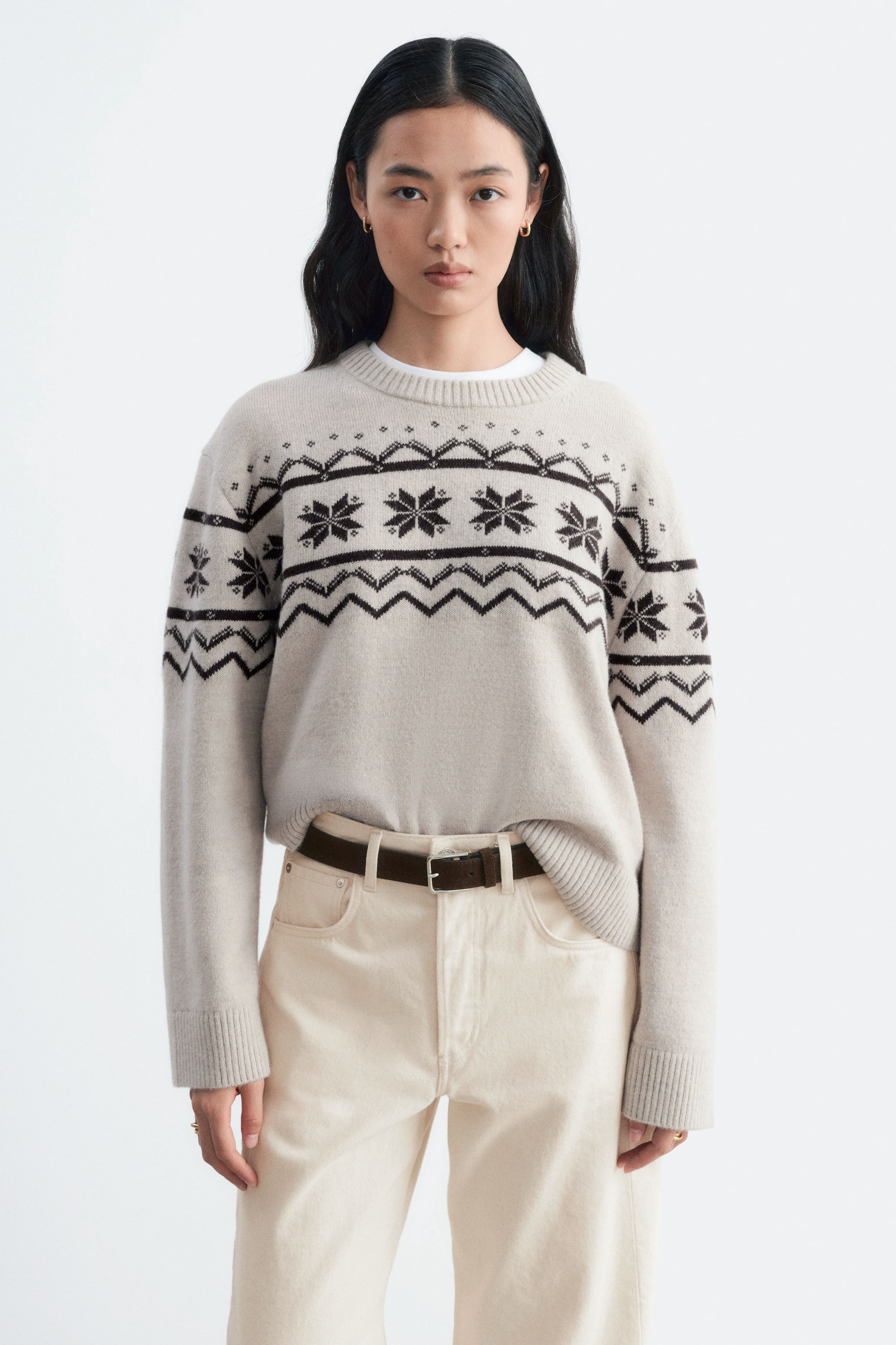 View larger image: A light beige knit jumper featuring a dark brown Fair Isle pattern across the chest, shoulders, and sleeves is being worn by the model. The jumper has a ribbed crew neck, cuffs, and hem, and is styled tucked into cream-coloured trousers with a brown belt.