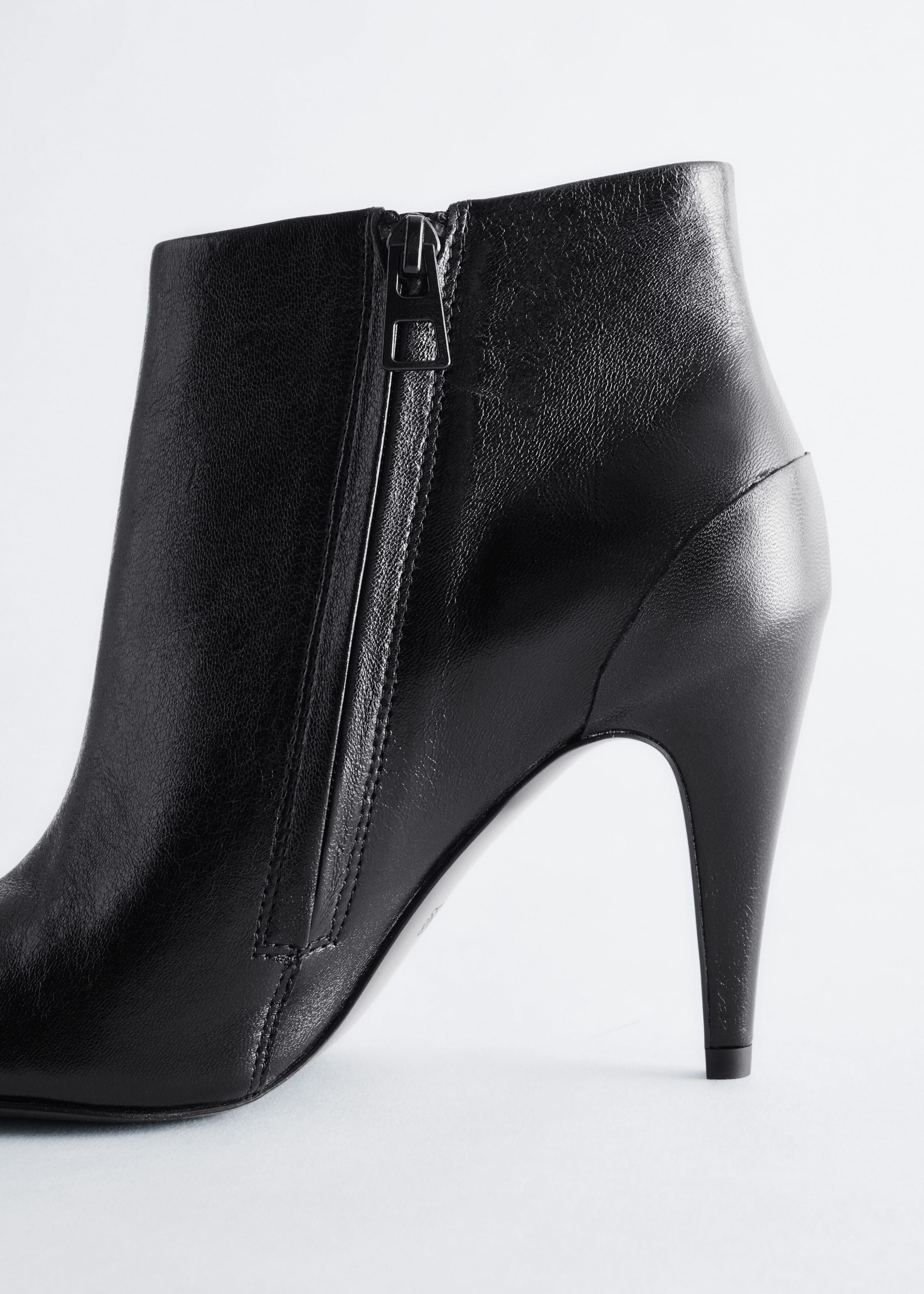 A black leather ankle boot is standing in a close-up side view facing left, showcasing its high stiletto heel and a side zip with a dark metal pull tab.