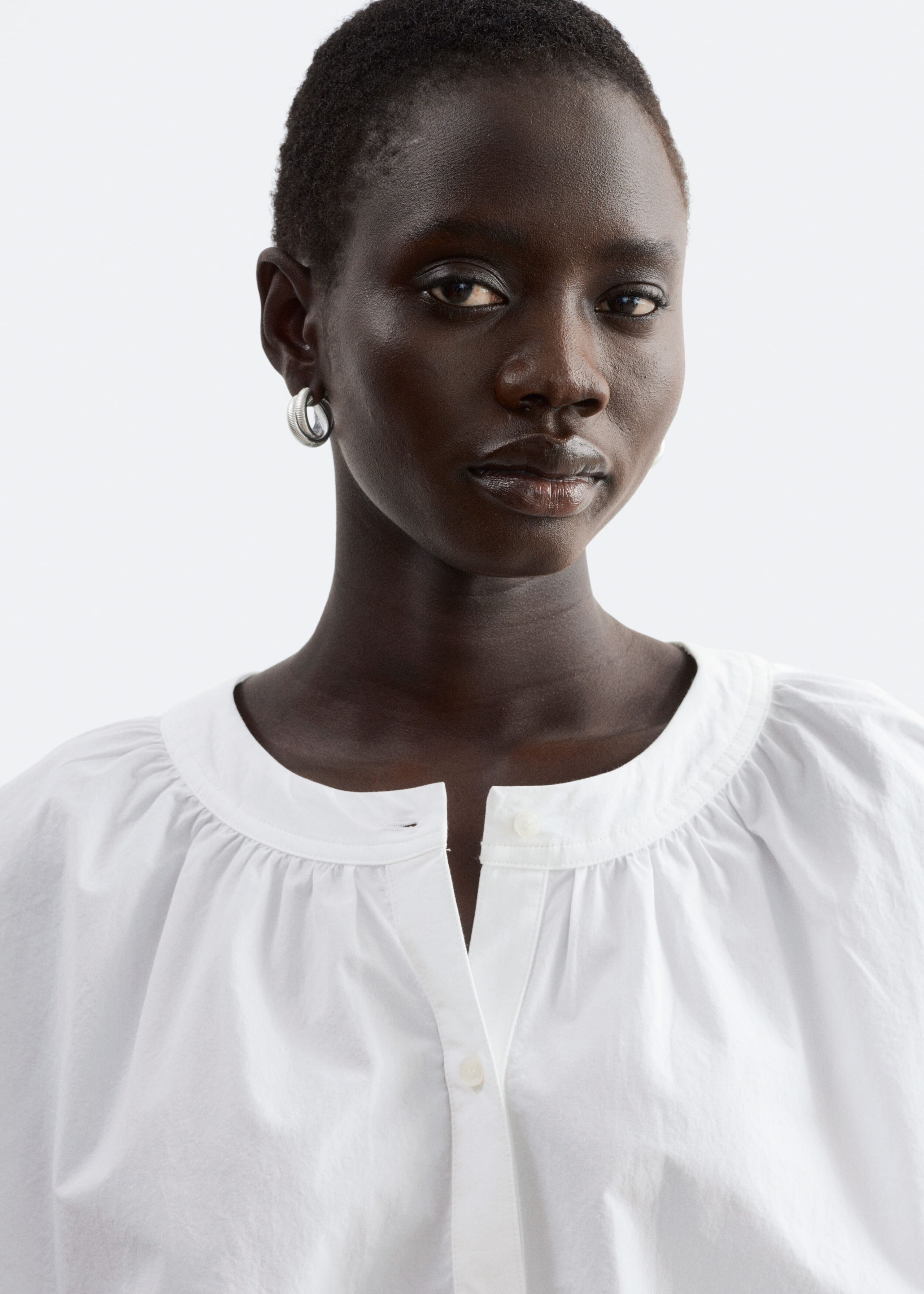 A white cotton blouse featuring a gathered neckline and a partially buttoned placket is being worn by the model. The close-up shot highlights the soft gathers that create volume around the neckline and shoulders.