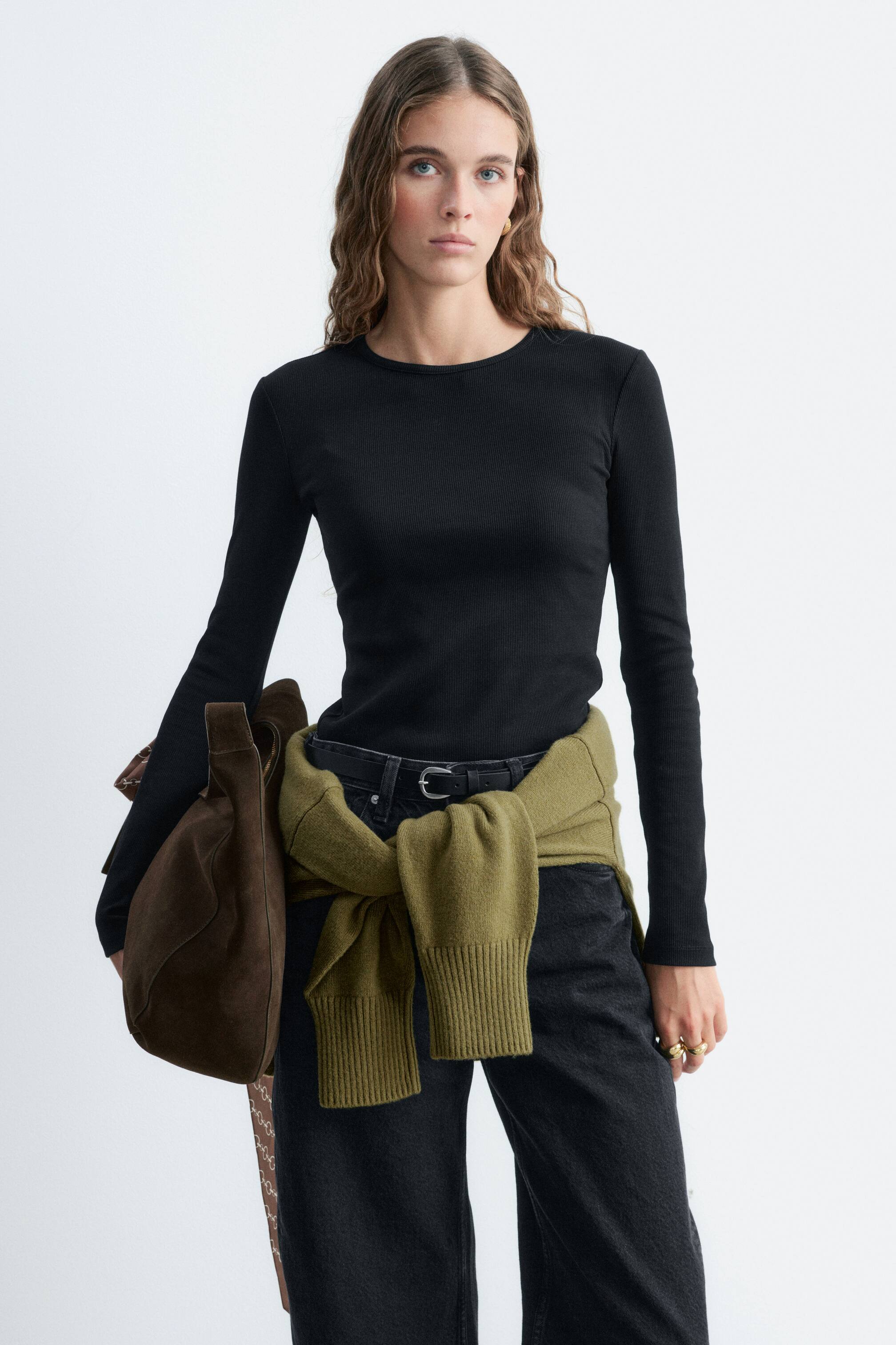 Grösseres Bild ansehen: Ein schwarzes Langarm-Oberteil mit Rippenstruktur, Rundhalsausschnitt und schmaler Passform wird vom Model getragen. Das Oberteil ist in dunkel gewaschene Jeans gesteckt und mit einem olivgrünen Strickpullover kombiniert, der um die Taille gebunden ist, sowie einer braunen Schultertasche aus Wildleder, die unter dem Arm getragen wird.