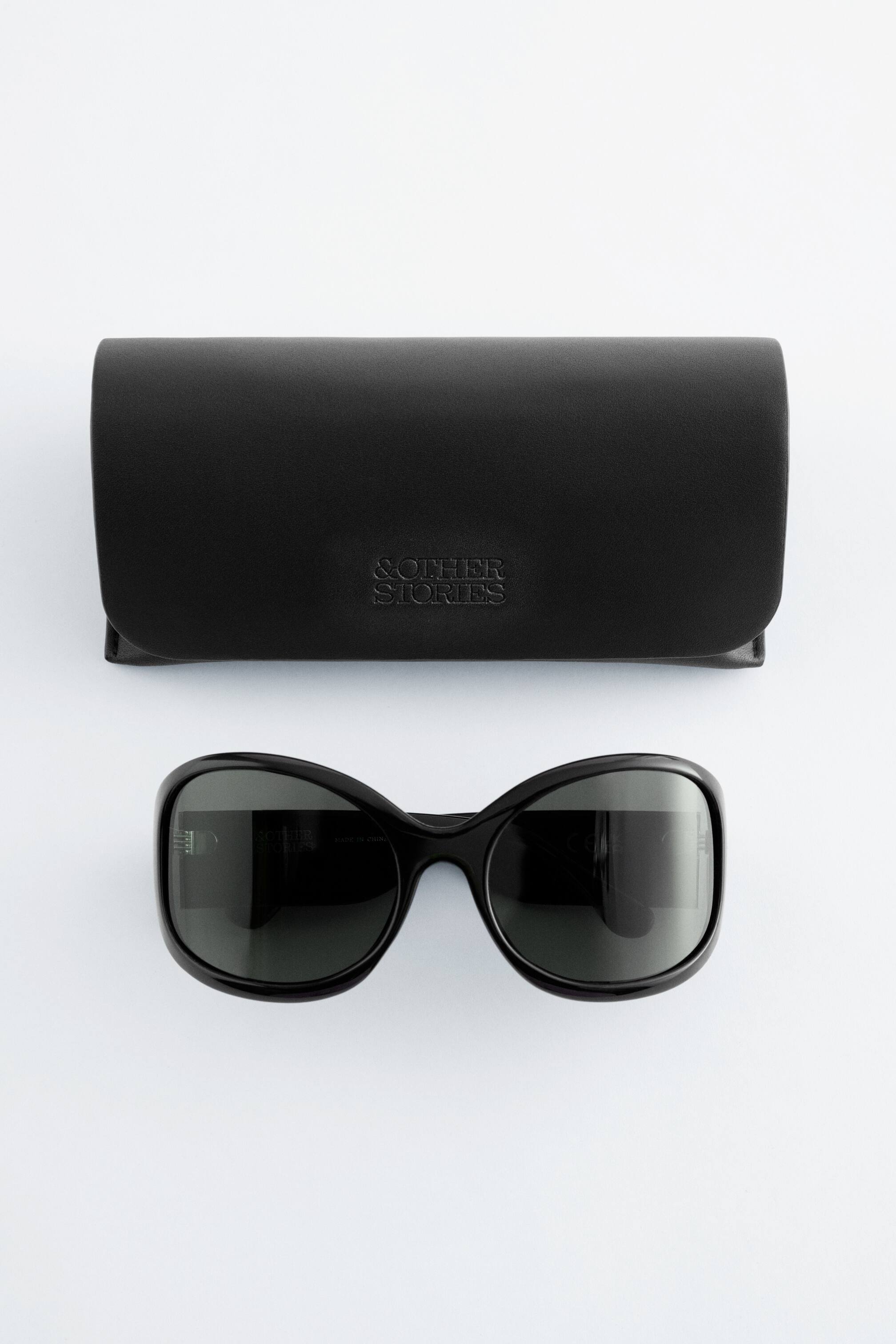 Agrandir l'image: Une paire de lunettes de soleil carrées surdimensionnées noires avec des montures légèrement arrondies, des verres teintés et des branches larges est présentée en vue de face. Un étui rectangulaire noir à parois souples, estampé de "& OTHER STORIES," est placé derrière les lunettes de soleil.