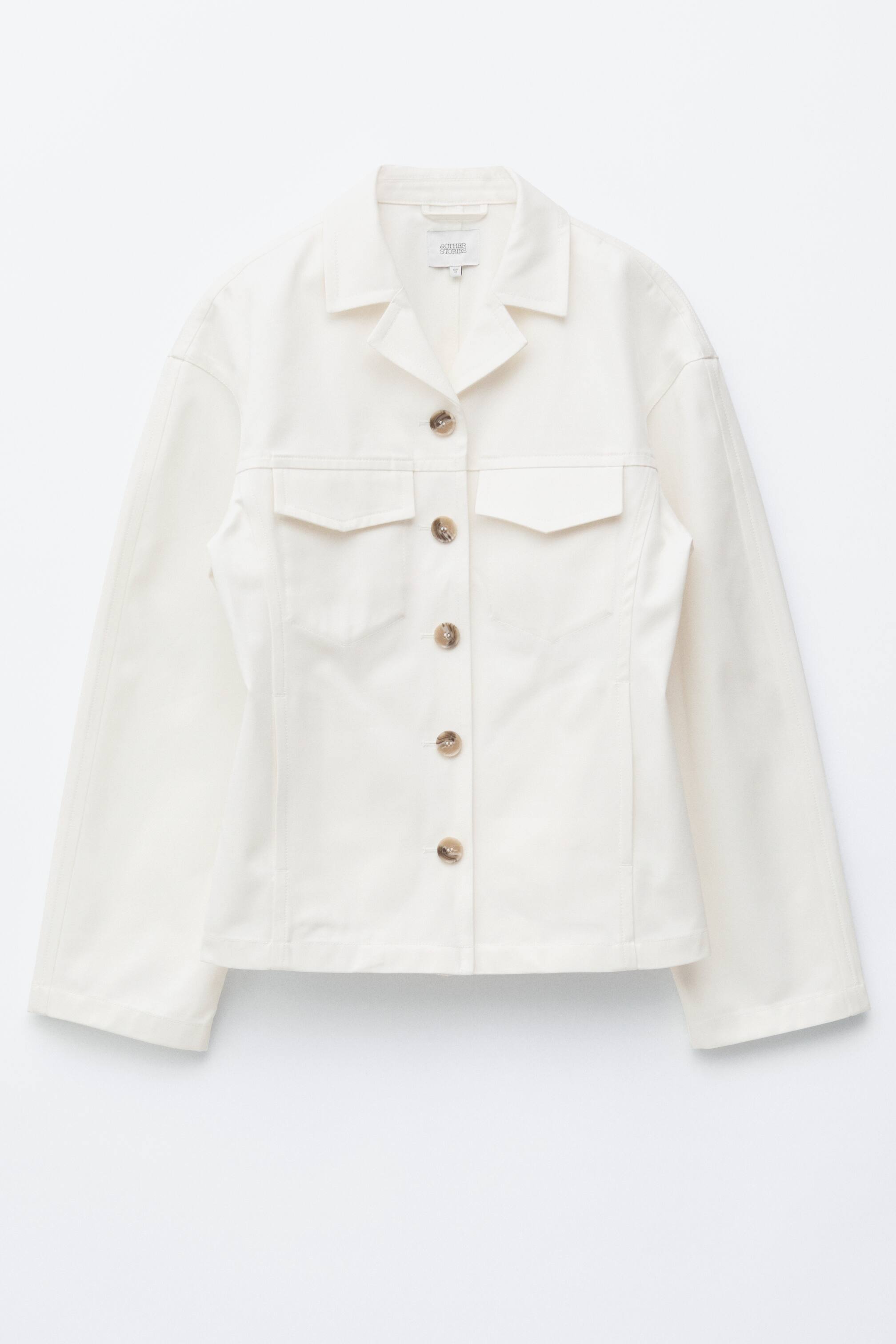 Ver imagem maior: Este casaco de sarja de algodão em tom off-white apresenta uma silhueta slim e de abotoamento simples, com a cintura ajustada na parte frontal. Destaca-se pelas lapelas em bico, dois bolsos de remendo com abas no peito e um fecho frontal de cinco botões com botões marmorizados.