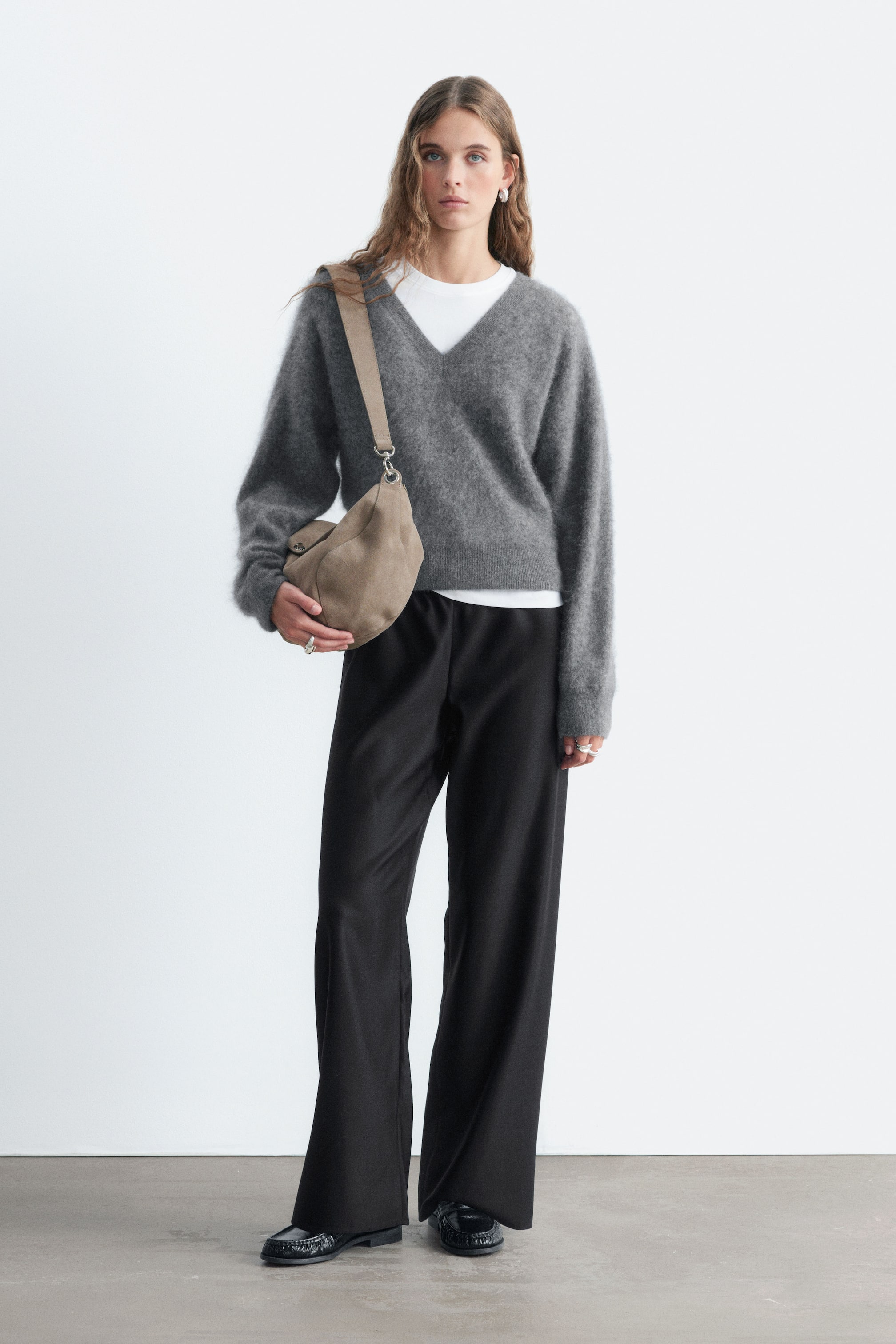 Grotere afbeelding bekijken: Zwarte instapbroeken van wol met een vloeiend silhouet en wijde pijpen worden gedragen door het model. De broek is gestyled met een grijze V-hals trui, een wit T-shirt en zwarte loafers.