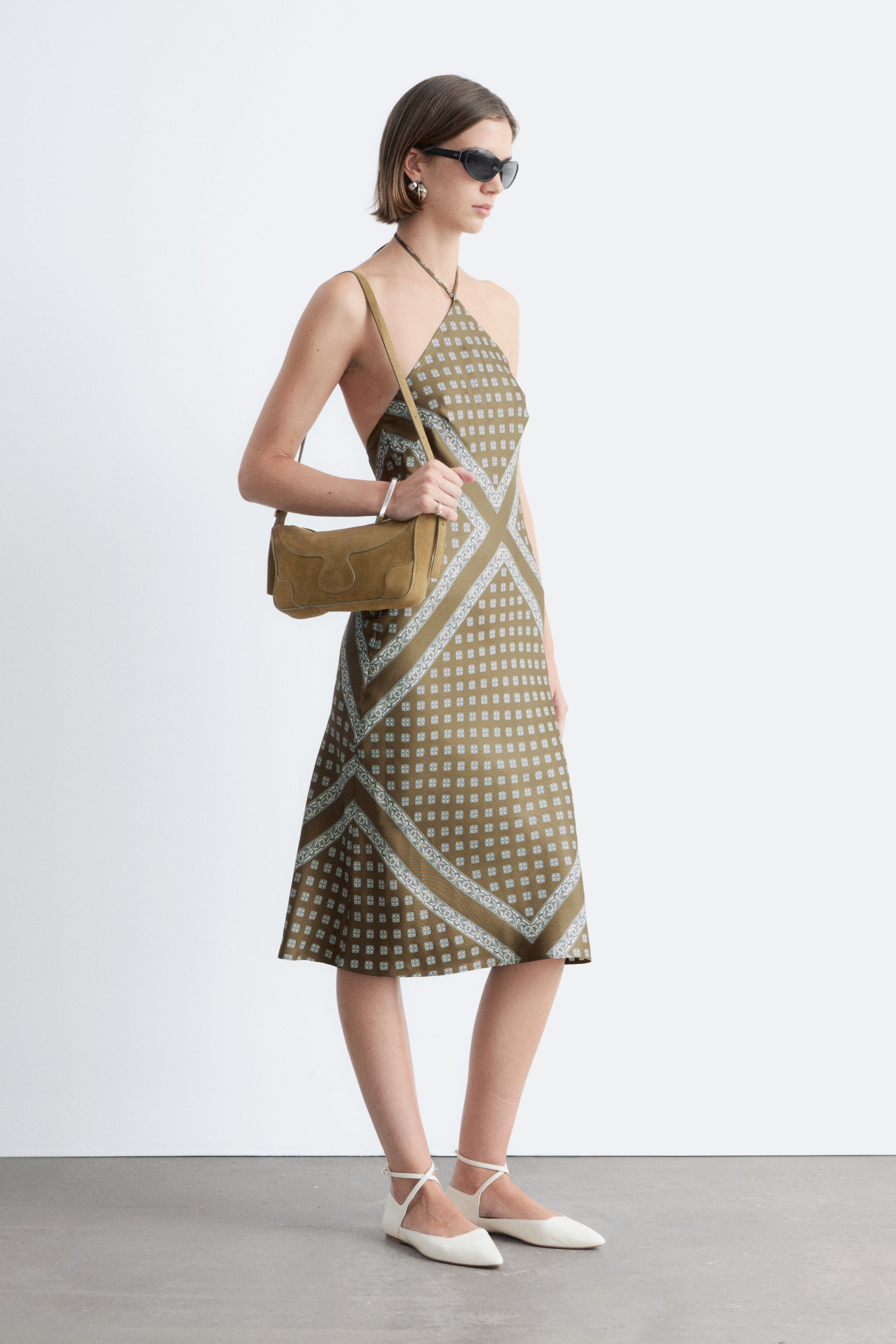 Se større billede: En olivengrøn halterneck-kjole med et geometrisk mønster bestående af små lyseblå kvadratmotiver og bredere diagonale bånd, der danner et gitter. Kjolen har en flydende A-formet silhuet, der strækker sig til lige under knæet.