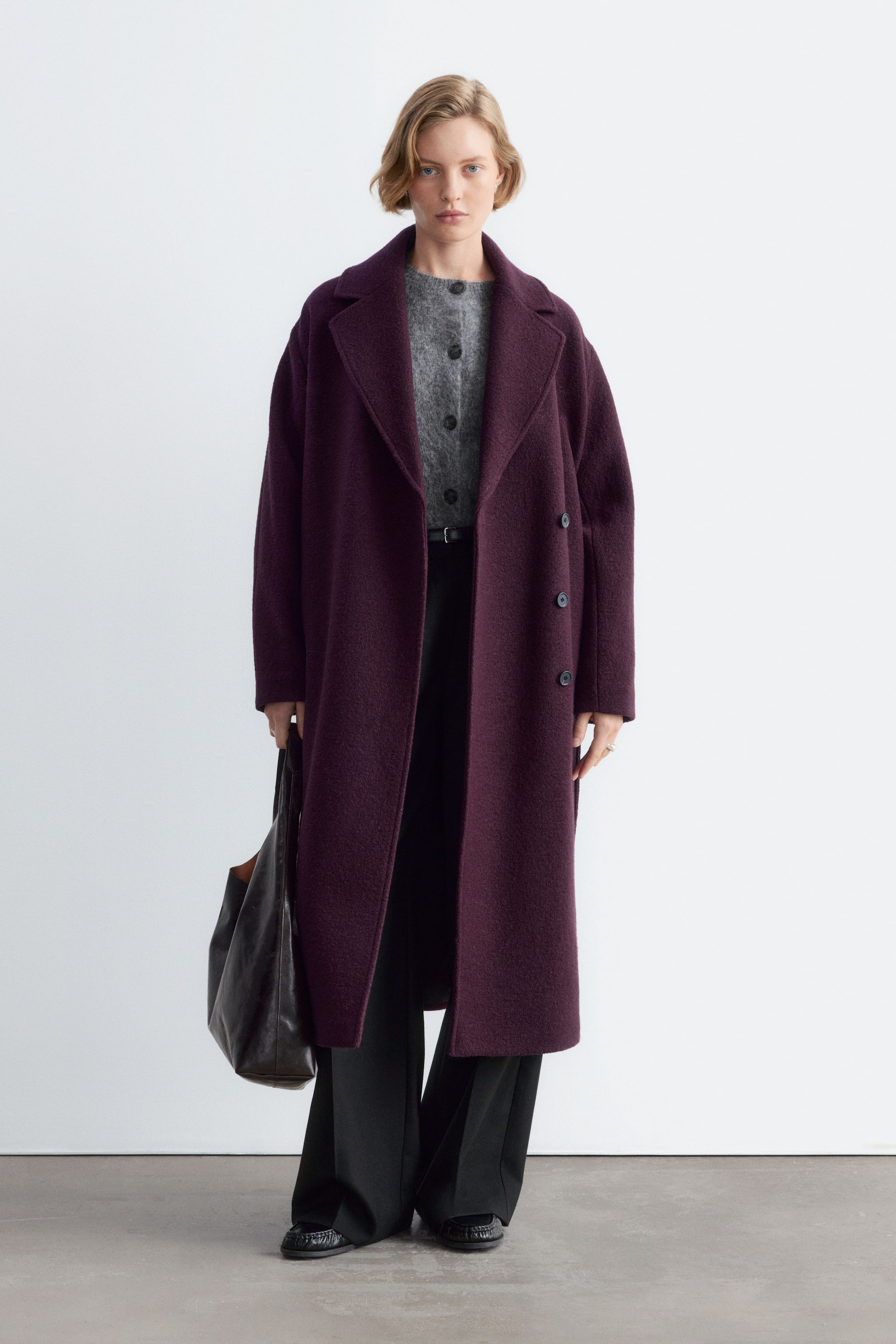 Visualizza l'immagine ingrandita: Un cappotto di lana viola intenso, doppiopetto e con ampi revers a lancia, è indossato aperto dalla modella. Il cappotto lungo è abbinato a un cardigan grigio abbottonato e a pantaloni neri, mentre viene portata una grande shopper in pelle marrone scuro.