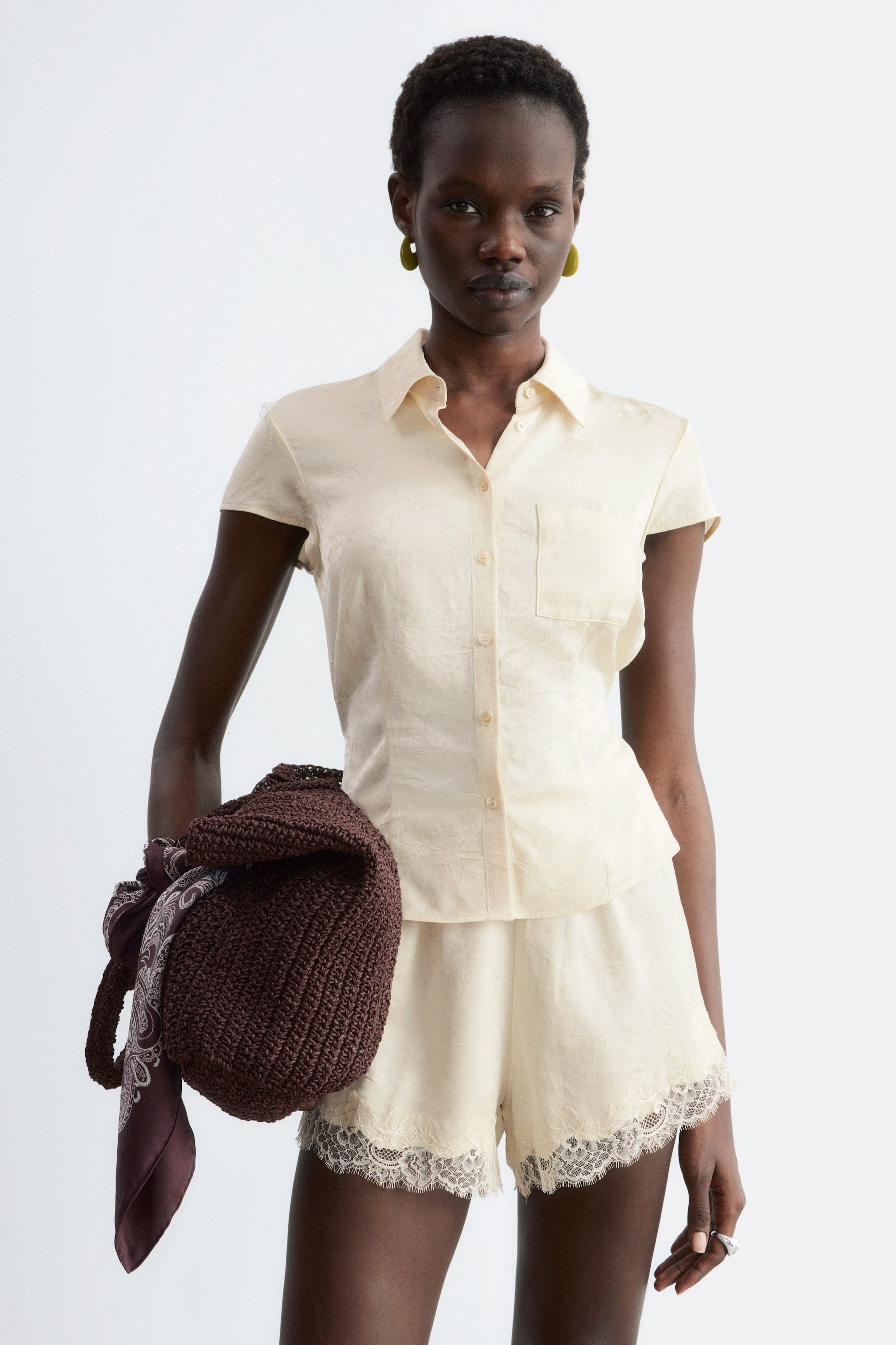 Visa större bild: En krämfärgad kortärmad skjorta med knappar och matchande shorts presenteras, båda med ett subtilt texturerat mönster. Skjortan har en spetsig krage, en knappslå och en enkel bröstficka på vänster sida, medan shortsen är designade med en vågig spetskant längs benöppningarna.