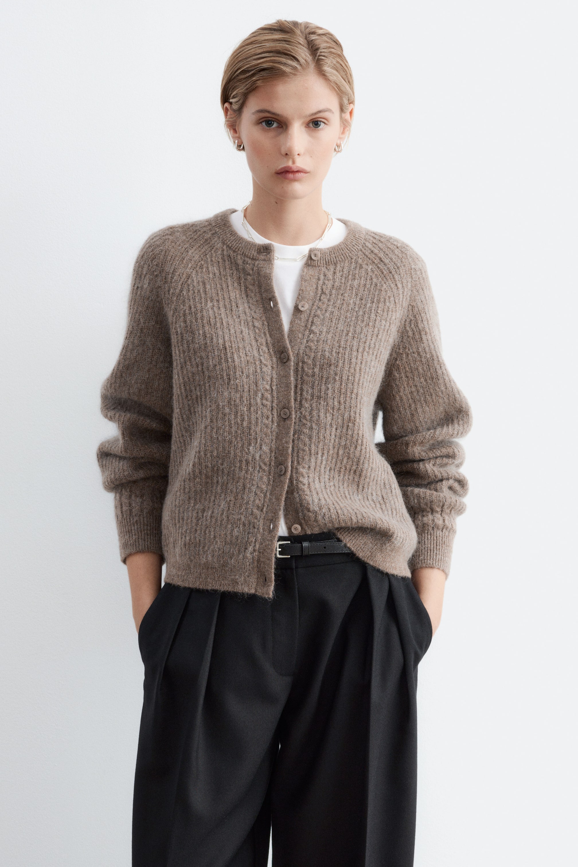 Agrandir l'image: Un cardigan marron en maille texturée, à manches raglan et fermeture boutonnée sur le devant, est porté par le mannequin. Le cardigan présente des détails torsadés le long de la patte de boutonnage et est superposé sur un t-shirt blanc, assorti à un pantalon noir.