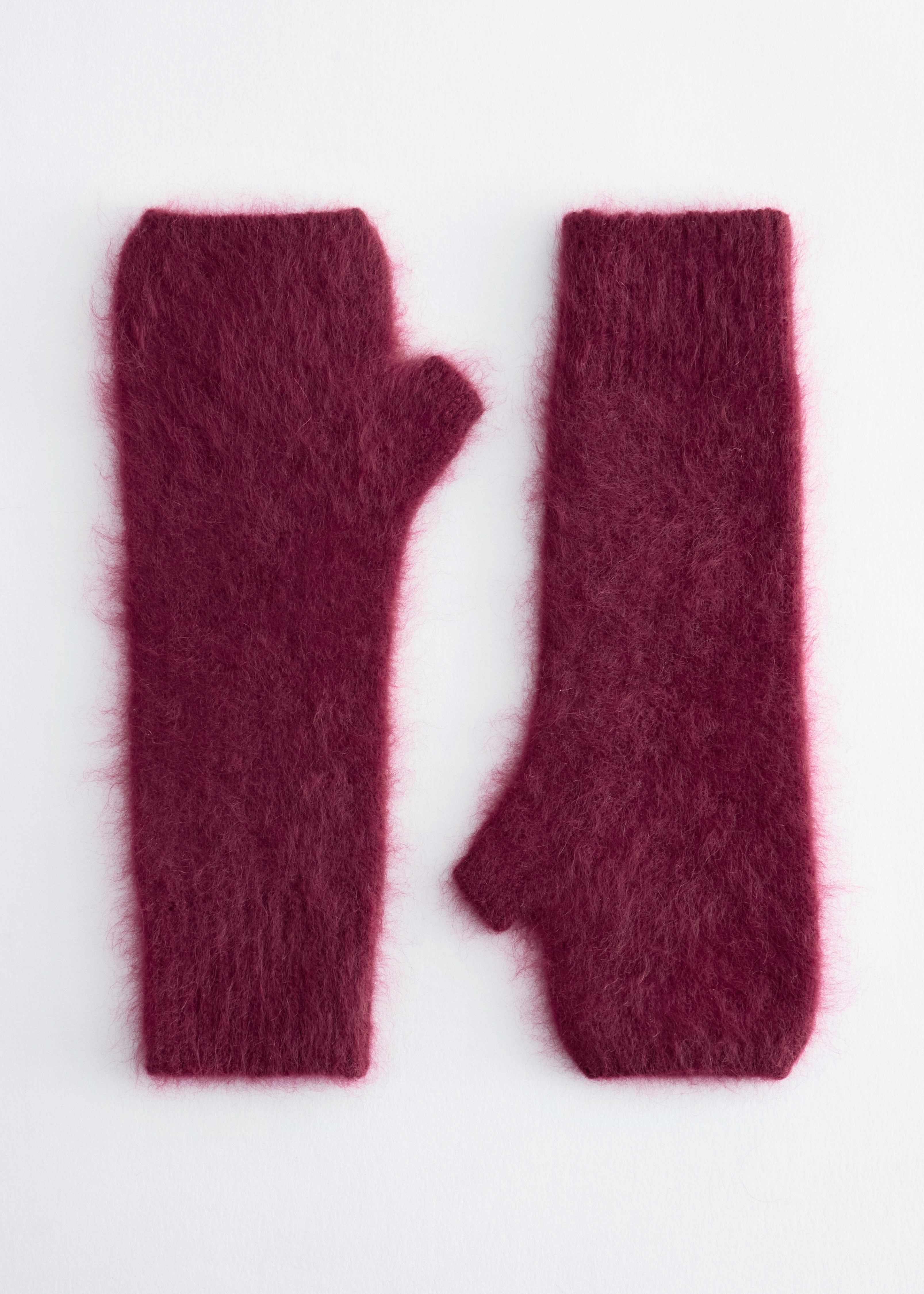 Burgundy fingerless gloves crafted from brushed cashmere are worn by the model, layered beneath the wide sleeves of a black coat. One gloved hand is raised towards the collar, highlighting the soft, fuzzy texture of the knitted accessory.