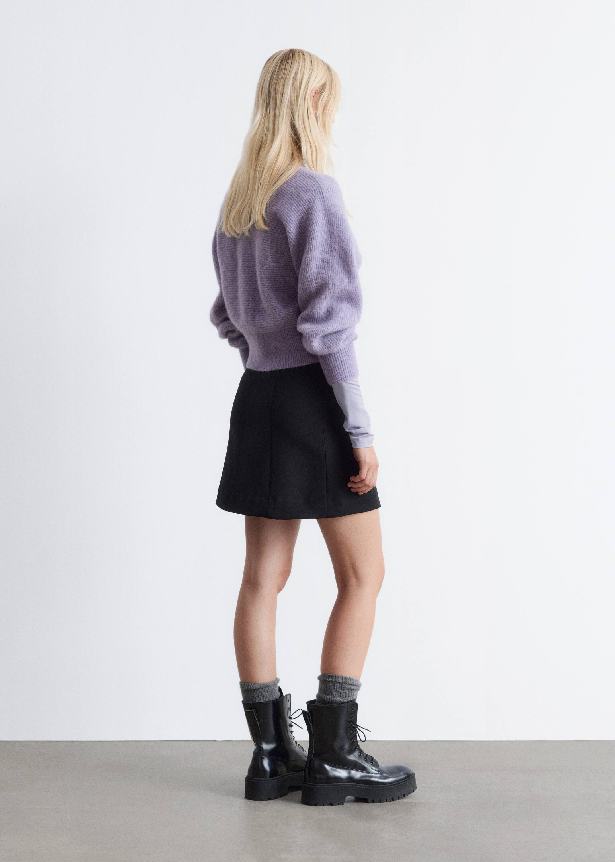 Agrandir l'image: Une tenue, présentée de l'arrière-droit, met en avant un pull court à manches longues de couleur violet clair, avec un ourlet froncé et des manches larges, superposé à un haut gris clair à manches longues s'étendant des poignets du pull. Celle-ci est associée à une jupe mini en ligne A noire et des bottines à lacets et semelles épaisses noires, avec des chaussettes grises côtelées visibles au-dessus des tiges des bottes.