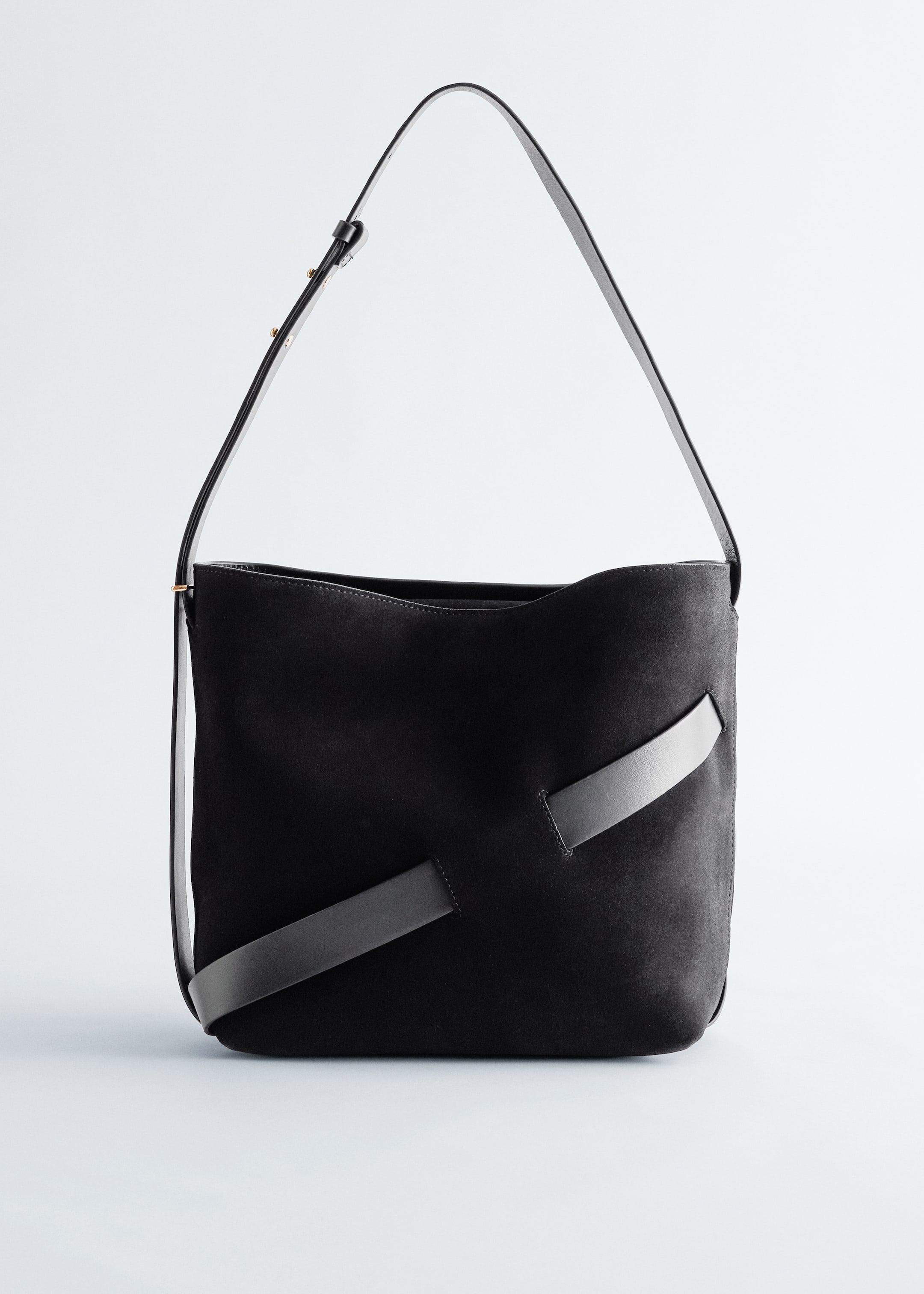 Größeres Bild anzeigen: Eine schwarze Wildleder-Bucket-Tasche wird von vorne präsentiert, mit einem verstellbaren schwarzen Lederschulterriemen mit sichtbaren goldfarbenen Beschlägen. Zwei zusätzliche schwarze Lederbesätze kreuzen diagonal die Vorderseite der Tasche.