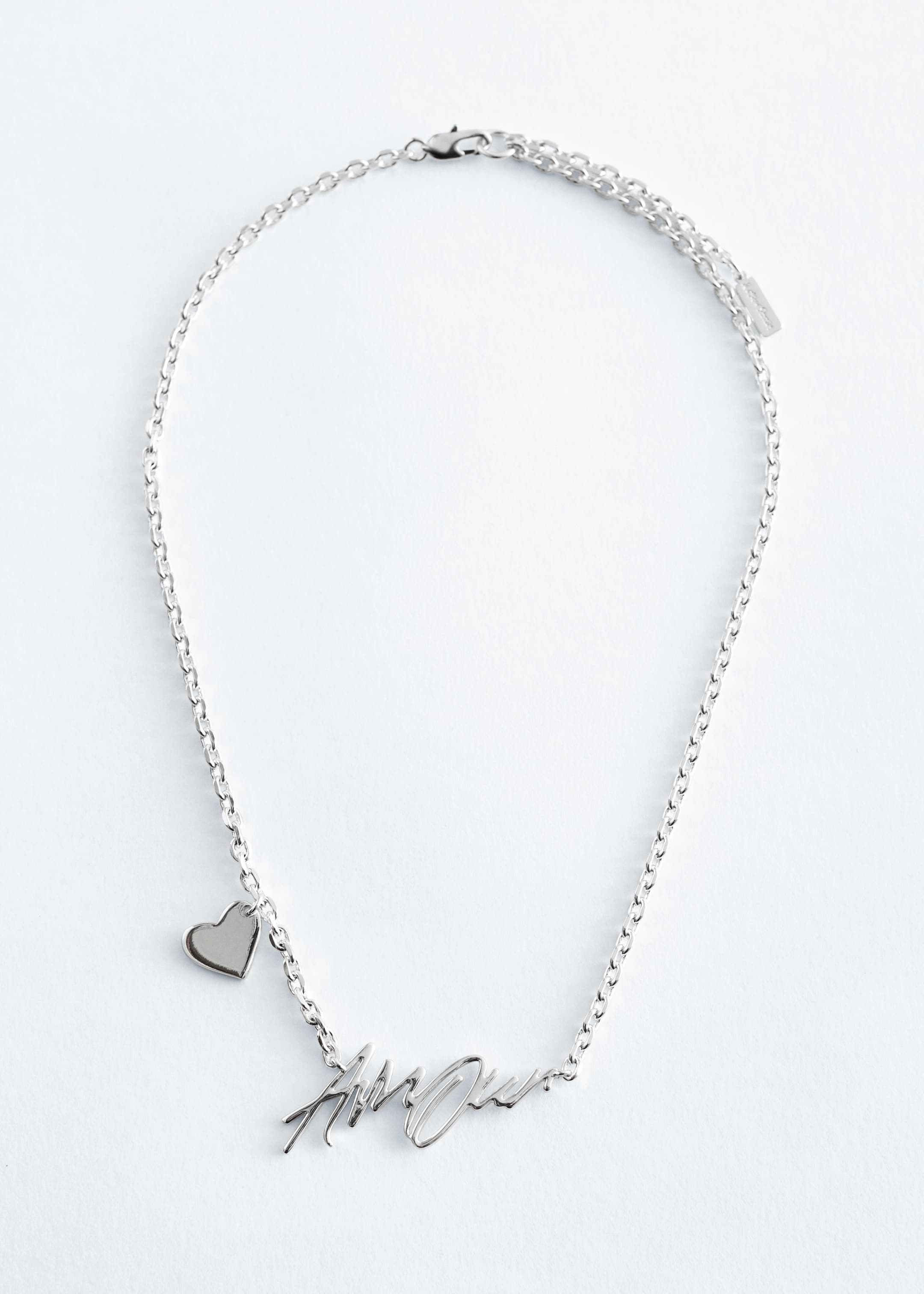 Grotere afbeelding bekijken: Een gepolijste, zilverkleurige ketting wordt gepresenteerd, met een delicate rolo-ketting en een hanger met tekstmotief die aan de voorkant 'Amour' spelt. Aan één kant van de ketting is een klein hartvormig bedeltje bevestigd, en de ketting is afgewerkt met een karabijnsluiting en een verstelbare lengte.