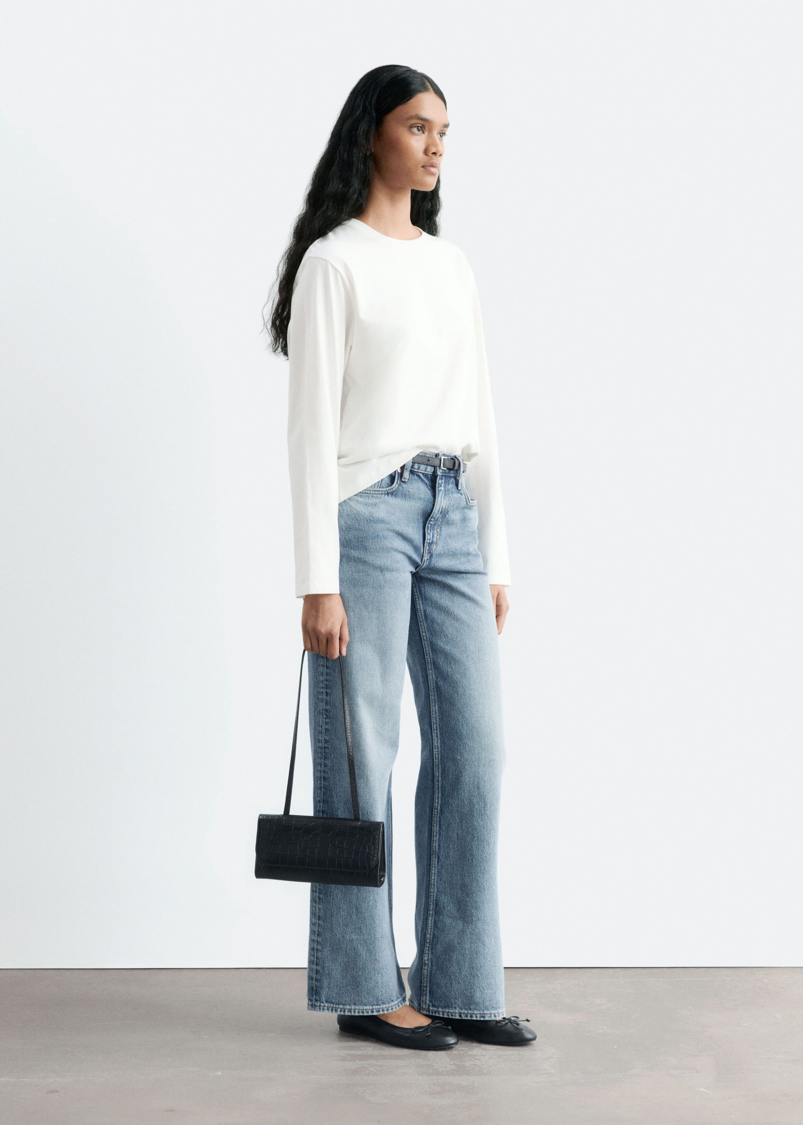 Light blue straight-leg denim jeans with a mid-rise waist are worn by the model, styled with a partially tucked-in white long-sleeve top and a slim black belt. The model is also wearing black ballet flats and holding a small black handbag.
