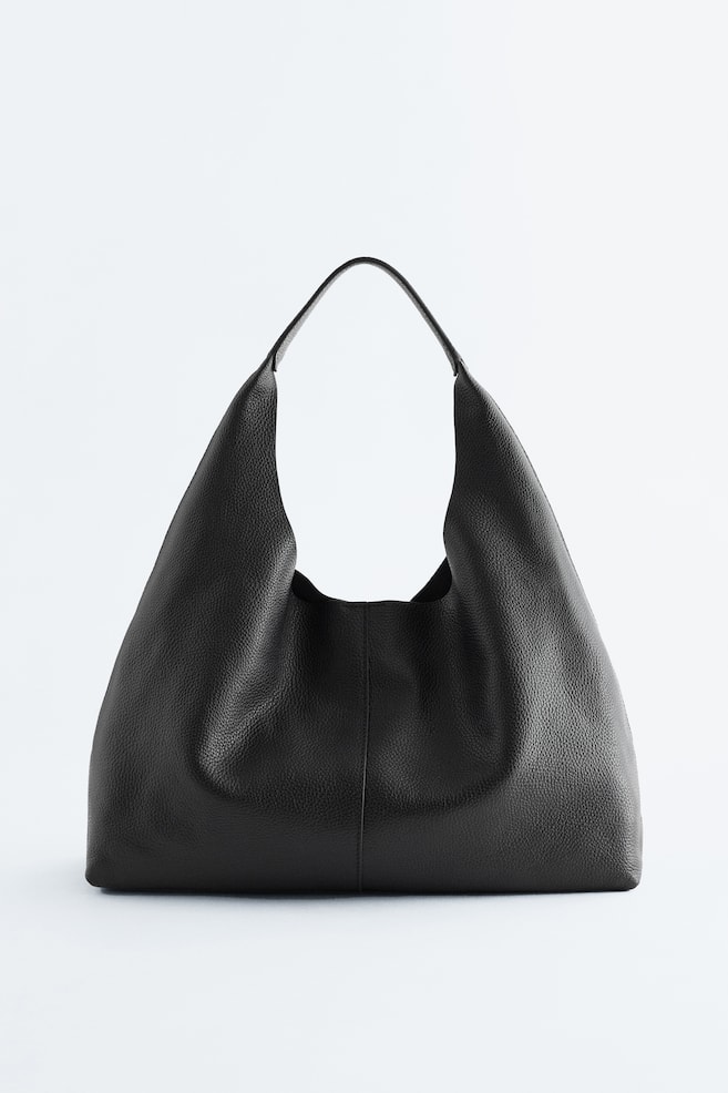 Una borsa tote nera è in piedi davanti, caratterizzata da una texture in pelle morbida e granulosa e un ampio manico integrato. Presenta una silhouette morbida con sottili cuciture verticali sul davanti. - 1
