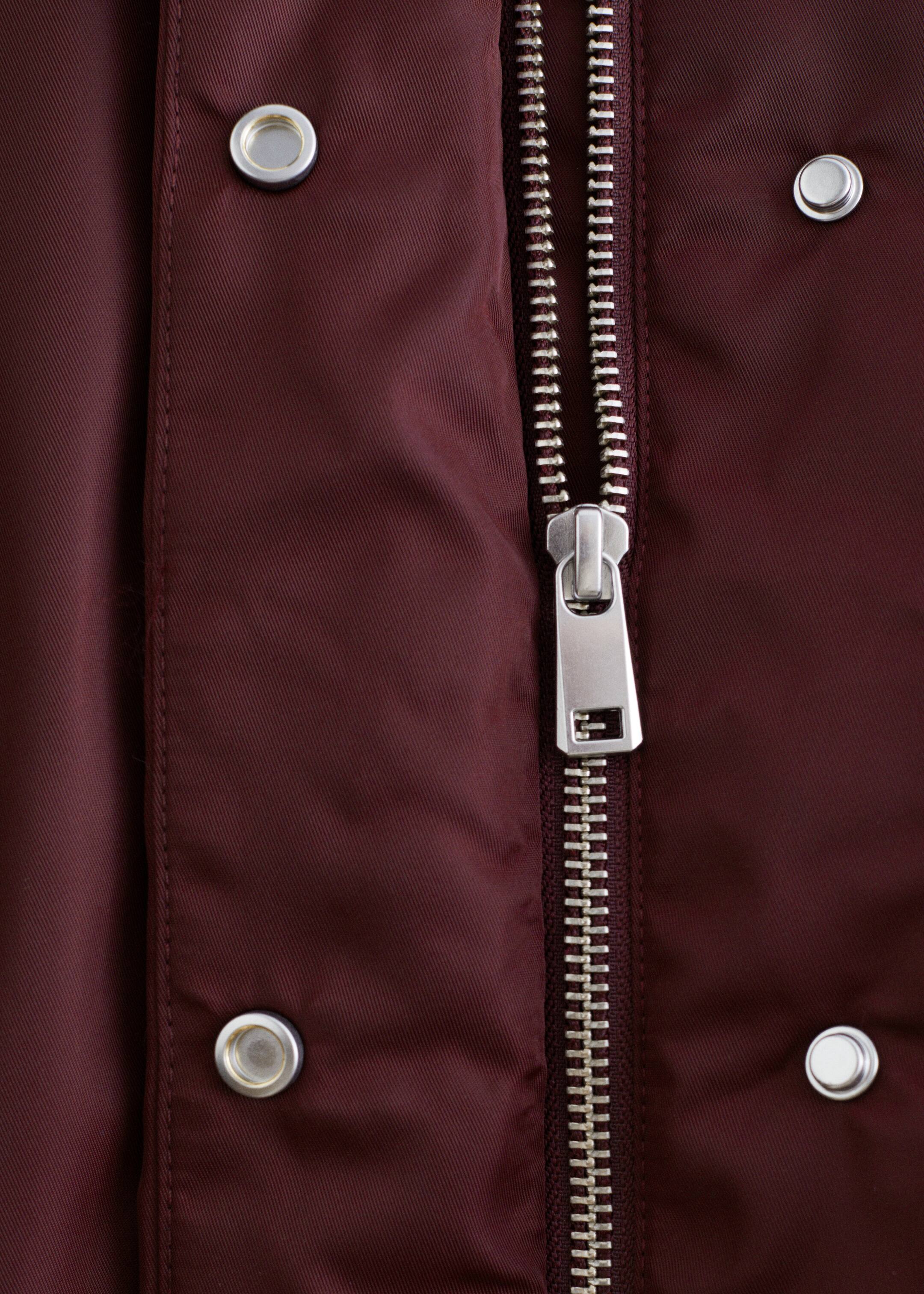 Visualizza immagine più grande: Una giacca bomber bordeaux, realizzata in un tessuto tecnico liscio, mostra la sua cerniera centrale in metallo color argento con un tirante rettangolare. I bottoni a pressione circolari color argento sono visibili sulla paramontura che fiancheggia la cerniera.