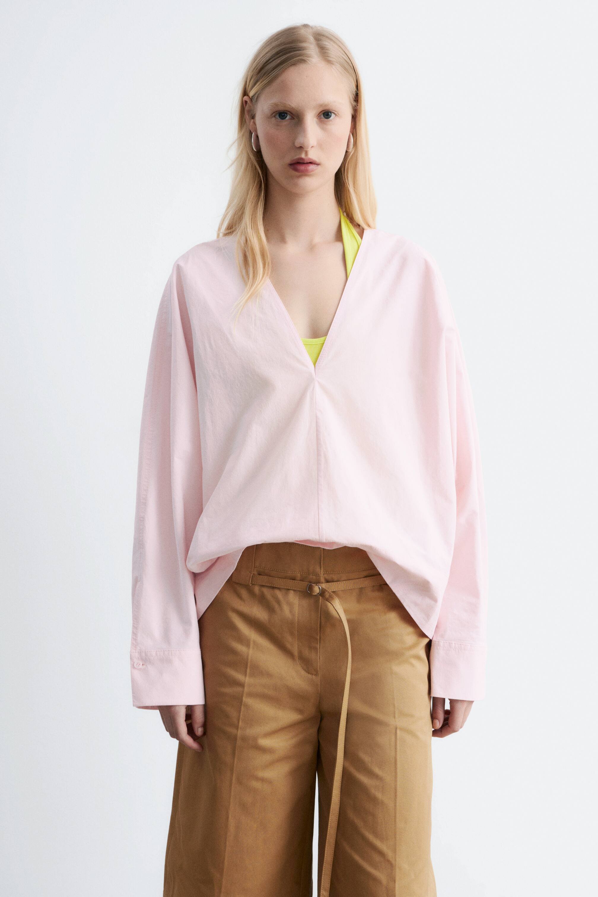 View larger image: A light pink V-neck blouse is being worn, featuring a relaxed fit, long sleeves with wide cuffs, and a deep V-neckline revealing a neon yellow top beneath.