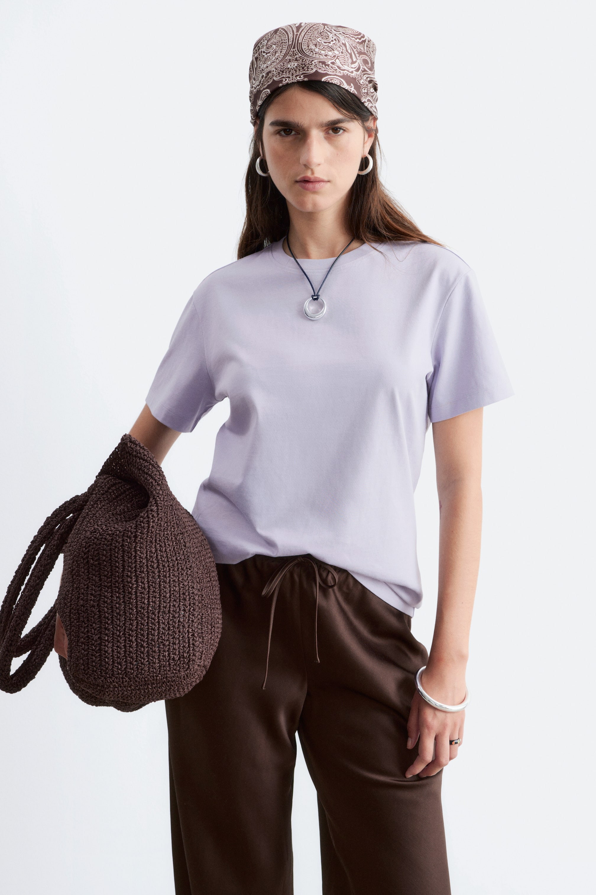 Zobacz większy obraz: Modelka ma na sobie jasnofioletowy bawełniany T-shirt z okrągłym dekoltem i krótkimi rękawami. Koszulka ma prosty, regularny krój, noszona jest na wierzchu brązowych spodni, a na niej znajduje się naszyjnik.