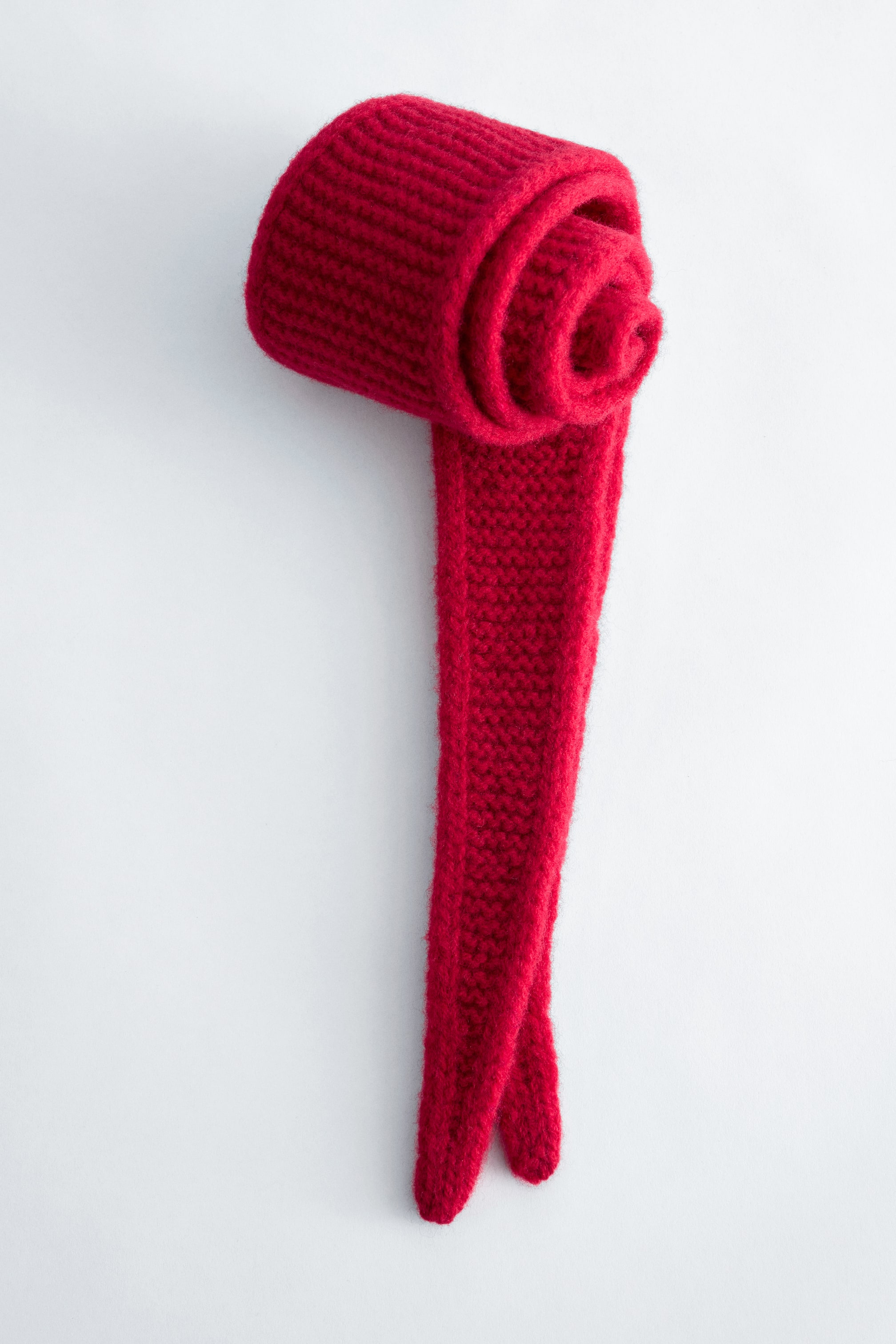 Agrandir l'image: Une écharpe en laine tricotée rouge est disposée avec une extrémité soigneusement enroulée en forme cylindrique, tandis que le reste s'étend vers le bas, révélant une texture douce et des extrémités effilées.