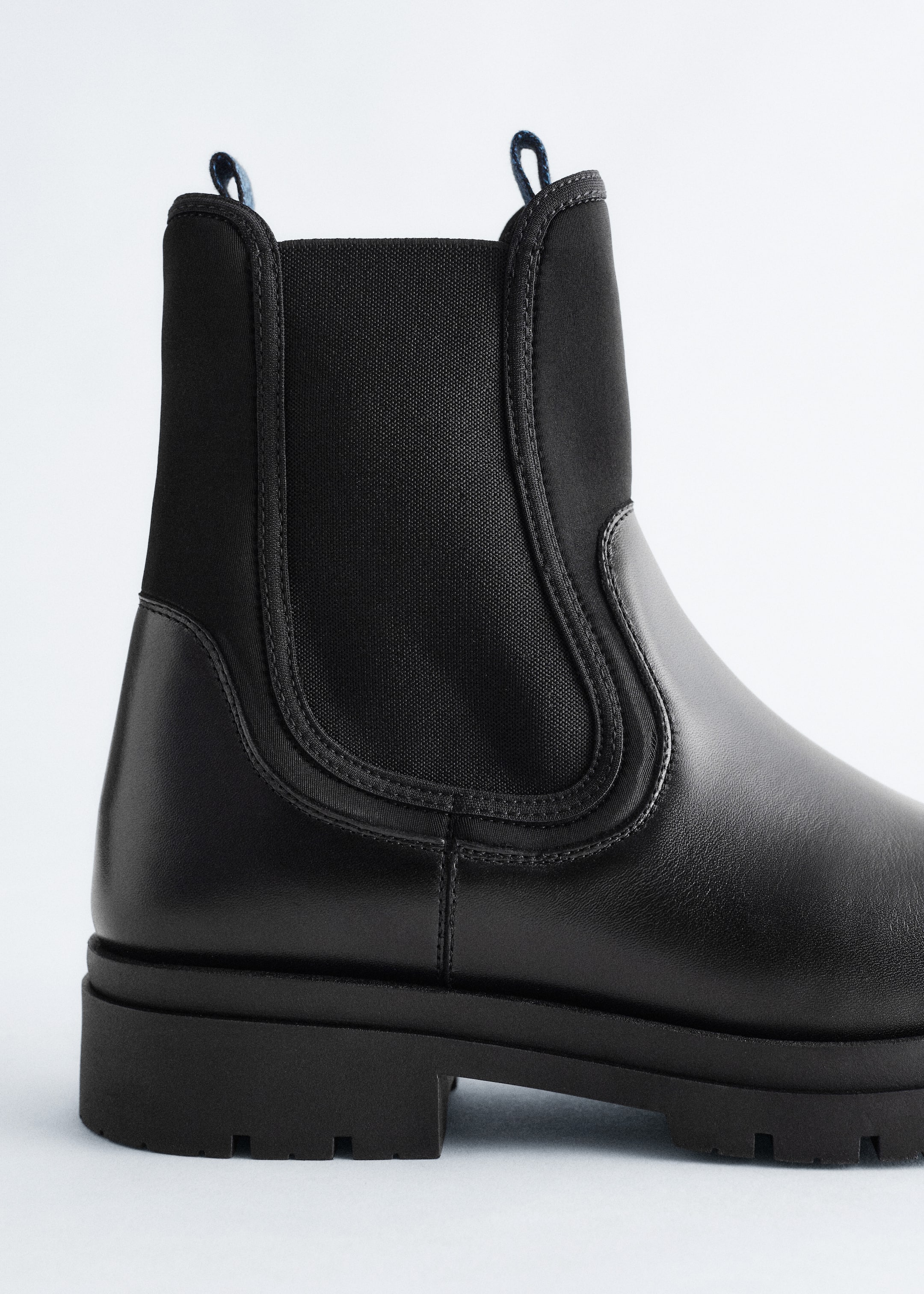 Ingrandisci l'immagine: Un Chelsea boot nero viene presentato da un'angolazione laterale, mettendo in mostra la tomaia e il tacco in pelle liscia, gli inserti elastici laterali e un gambale in scuba morbido con linguette blu nella parte superiore, il tutto su una suola robusta e dentellata.