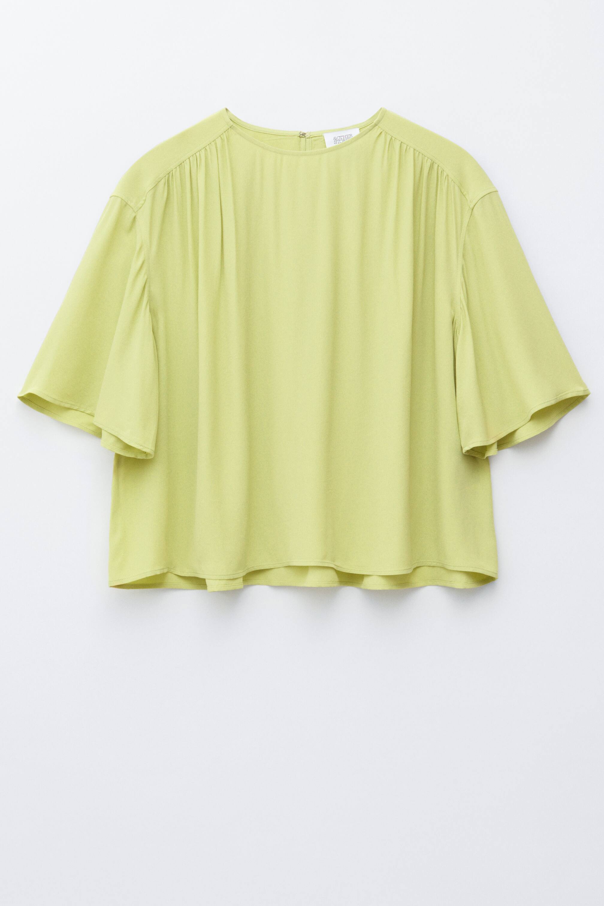 Agrandir l'image: Un haut vert citron doté d'un col rond et de manches évasées trois-quarts. Il présente des détails froncés le long des coutures des épaules, créant une coupe décontractée et drapée, avec une petite fermeture éclair visible à l'arrière du col.