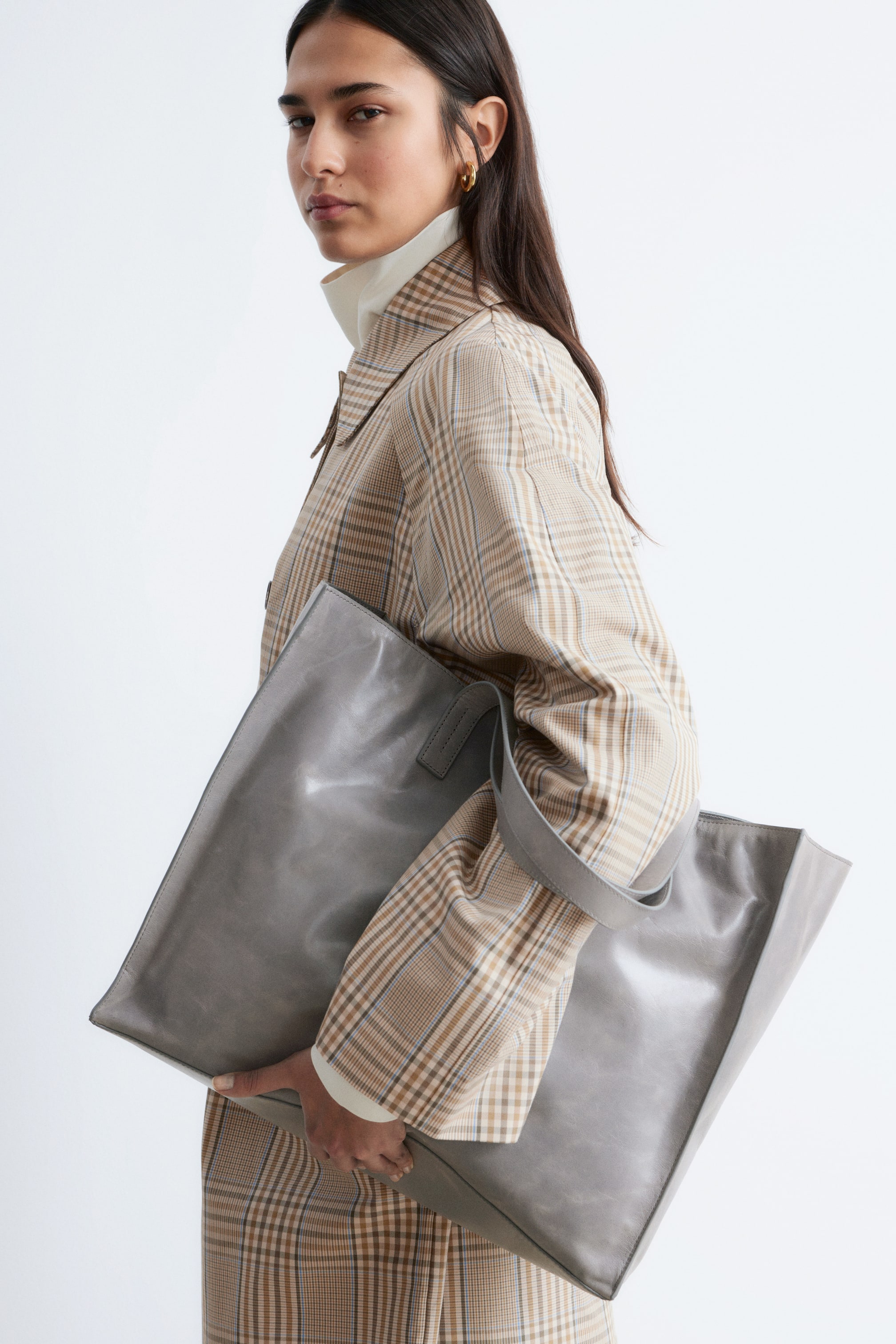 Se større billede: En stor, grå læder tote taske med en minimalistisk silhuet og to top håndtag bæres af modellen. Modellen holder tasken tæt til kroppen, som er klædt i en ternet jakke.