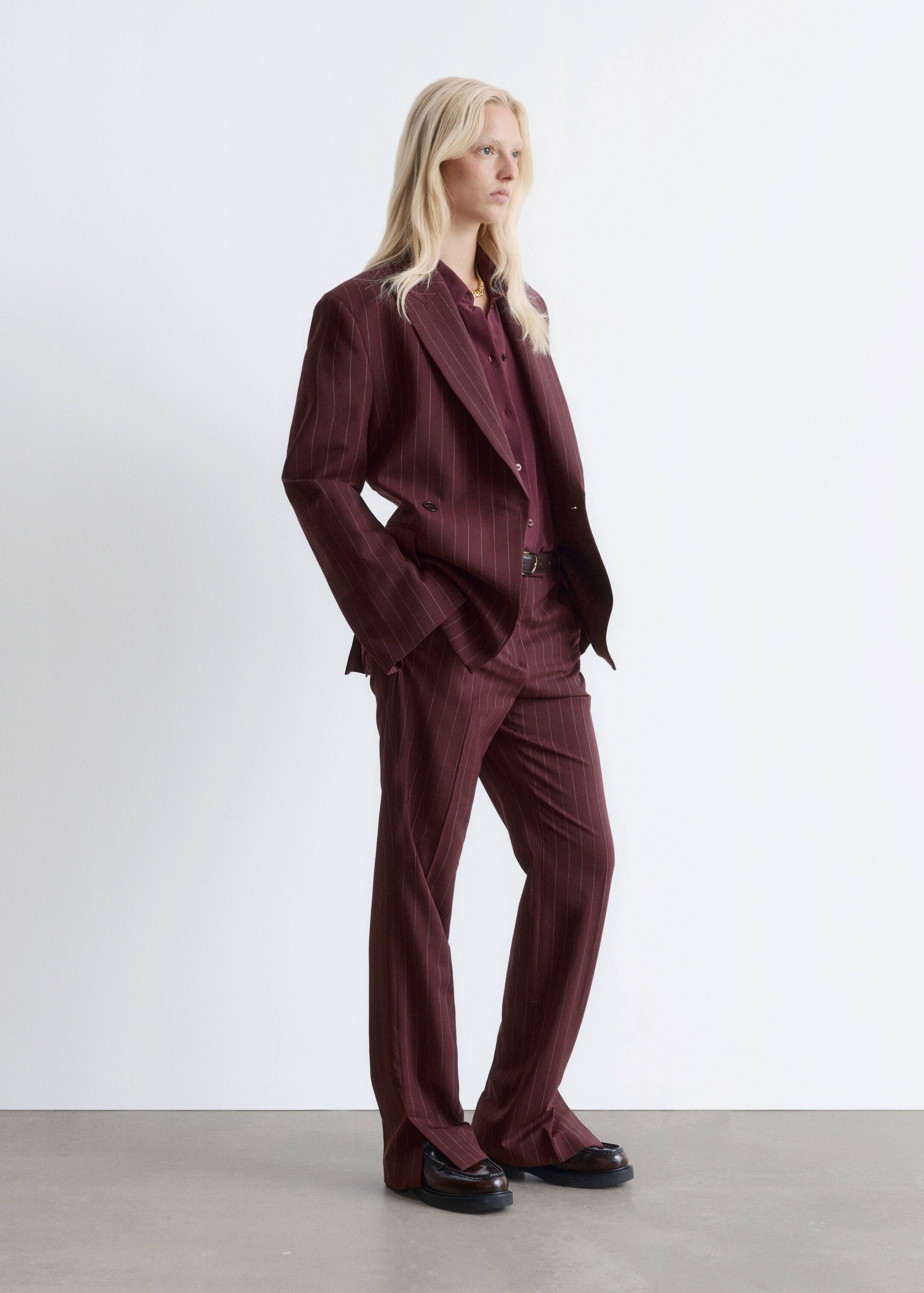 Agrandir l'image: Un costume à rayures bordeaux est présenté sous un angle légèrement incliné, mettant en valeur son blazer croisé oversize avec des revers en pointe et un pantalon large assorti. Une chemise bordeaux foncé est portée sous le blazer, complétant l'ensemble coordonné avec des mocassins sombres.