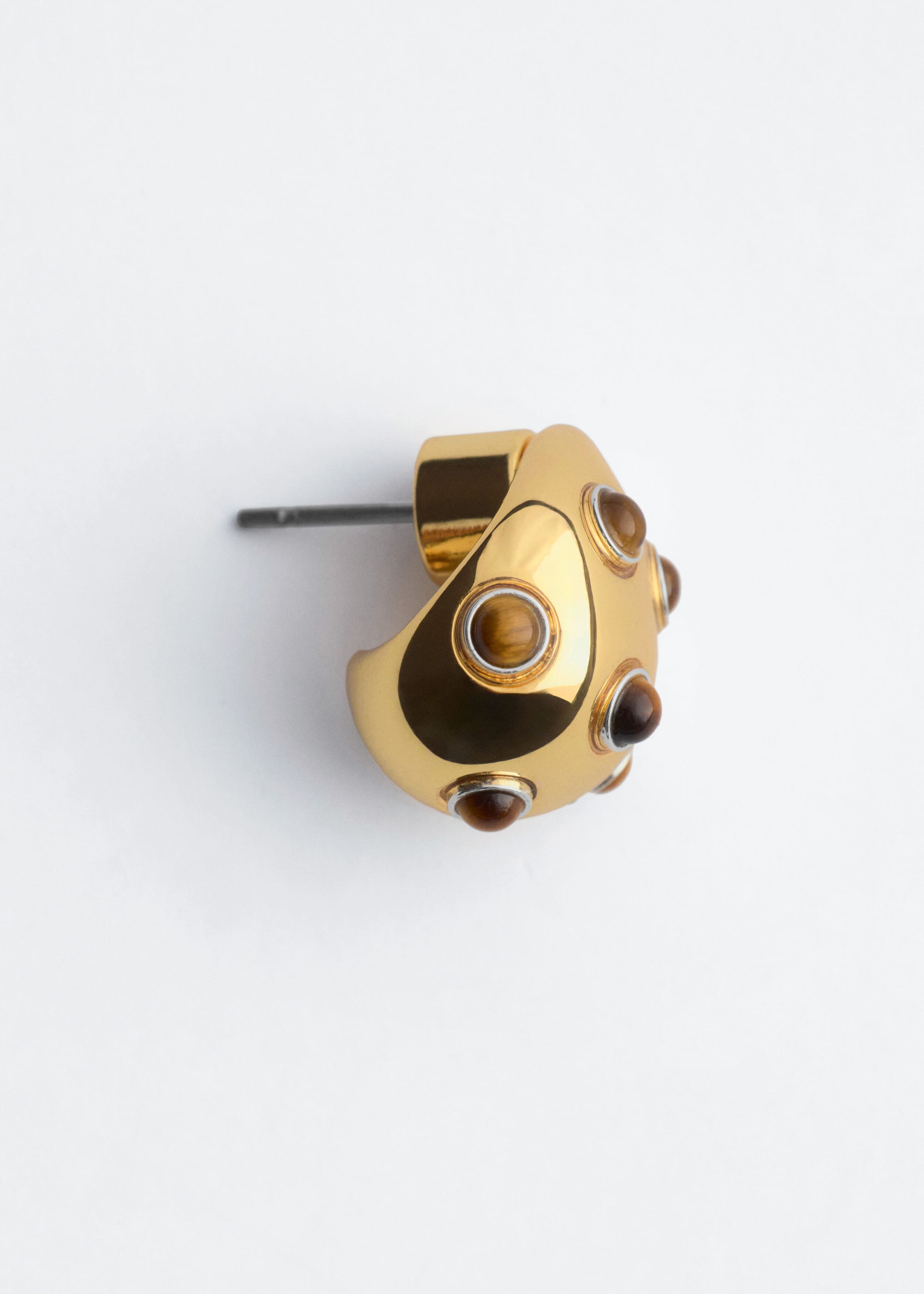 Un orecchino in ottone lucidato presenta una forma a cupola delicatamente arrotondata, impreziosita da pietre di occhio di tigre circolari incastonate in castoni. L’orecchino è orientato verso sinistra, mettendo in mostra il perno metallico dritto e il profilo curvo.