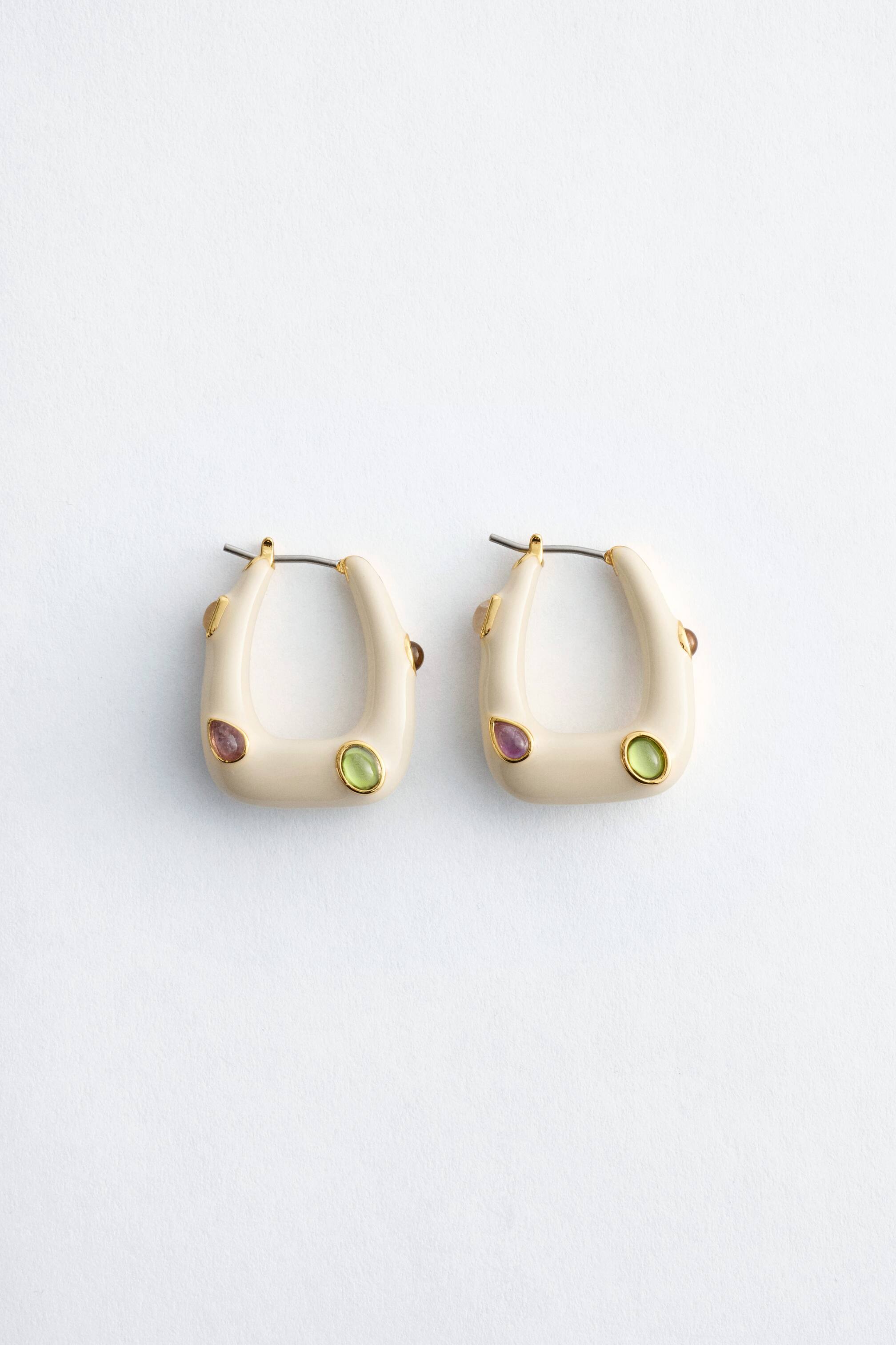 Agrandir l'image: Une paire de boucles d'oreilles en forme de U de couleur beige clair, ornée d'une pierre précieuse en forme de goutte d'eau violette encadrée d'or et d'une pierre précieuse ovale vert citron encadrée d'or sur le devant, ainsi qu'une plus petite pierre précieuse brune sur le bord extérieur.