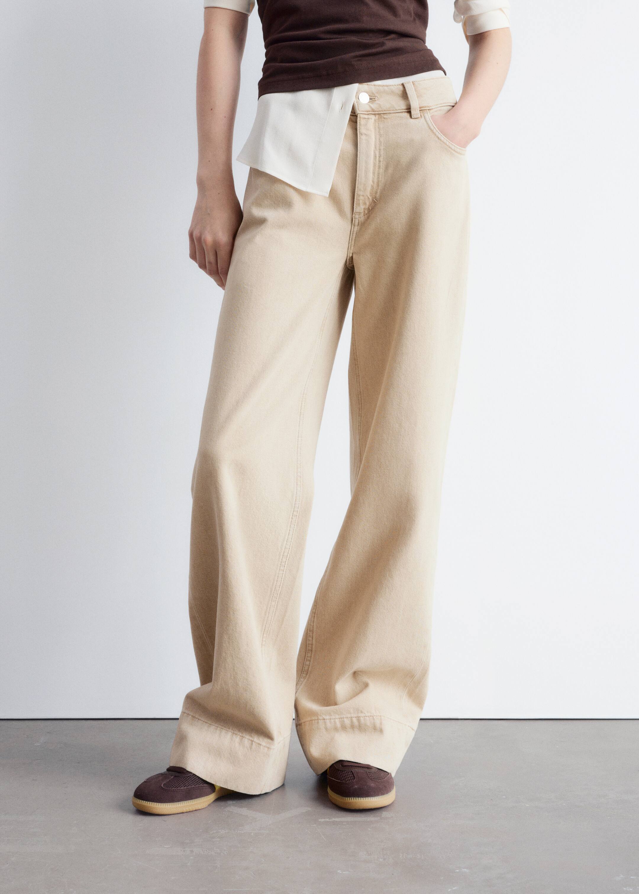 Größeres Bild anzeigen: Weite Hosen in hellem Beige werden präsentiert, mit hoher Taille, Gürtelschlaufen und Vordertaschen. Auffällige vertikale Ziernähte verlaufen entlang der Vorderseite jedes Beins, und die Hosen fallen über dunkelbraune Schuhe.