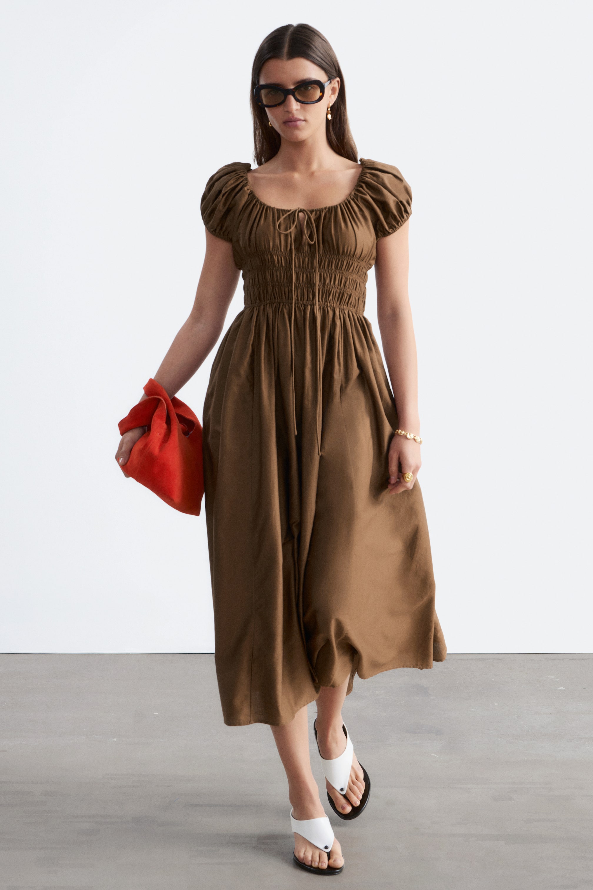 View larger image: The model is wearing a brown midi dress with puff sleeves, a smocked bodice, and a drawstring neckline. She complements the dress with an orange bag, sunglasses, and white sandals.