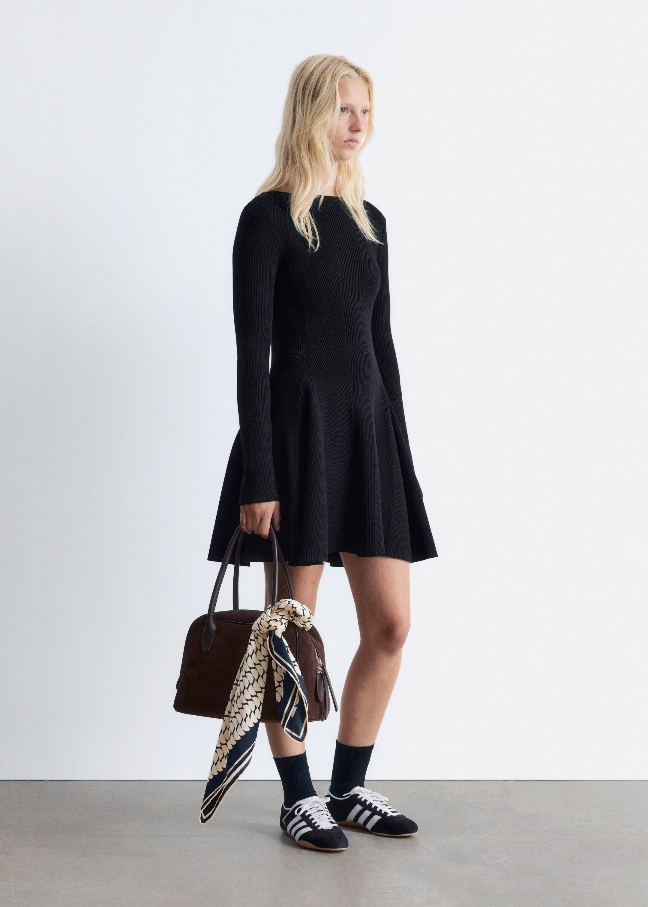 Ingrandisci l'immagine: Un mini abito nero, a coste, lavorato a maglia con un'ampia scollatura, vita aderente e gonna svasata è indossato dalla modella. Scarpe sportive scure con strisce bianche e lacci sono indossate con calzini scuri, e una borsa marrone con un foulard a fantasia legato al manico è portata.