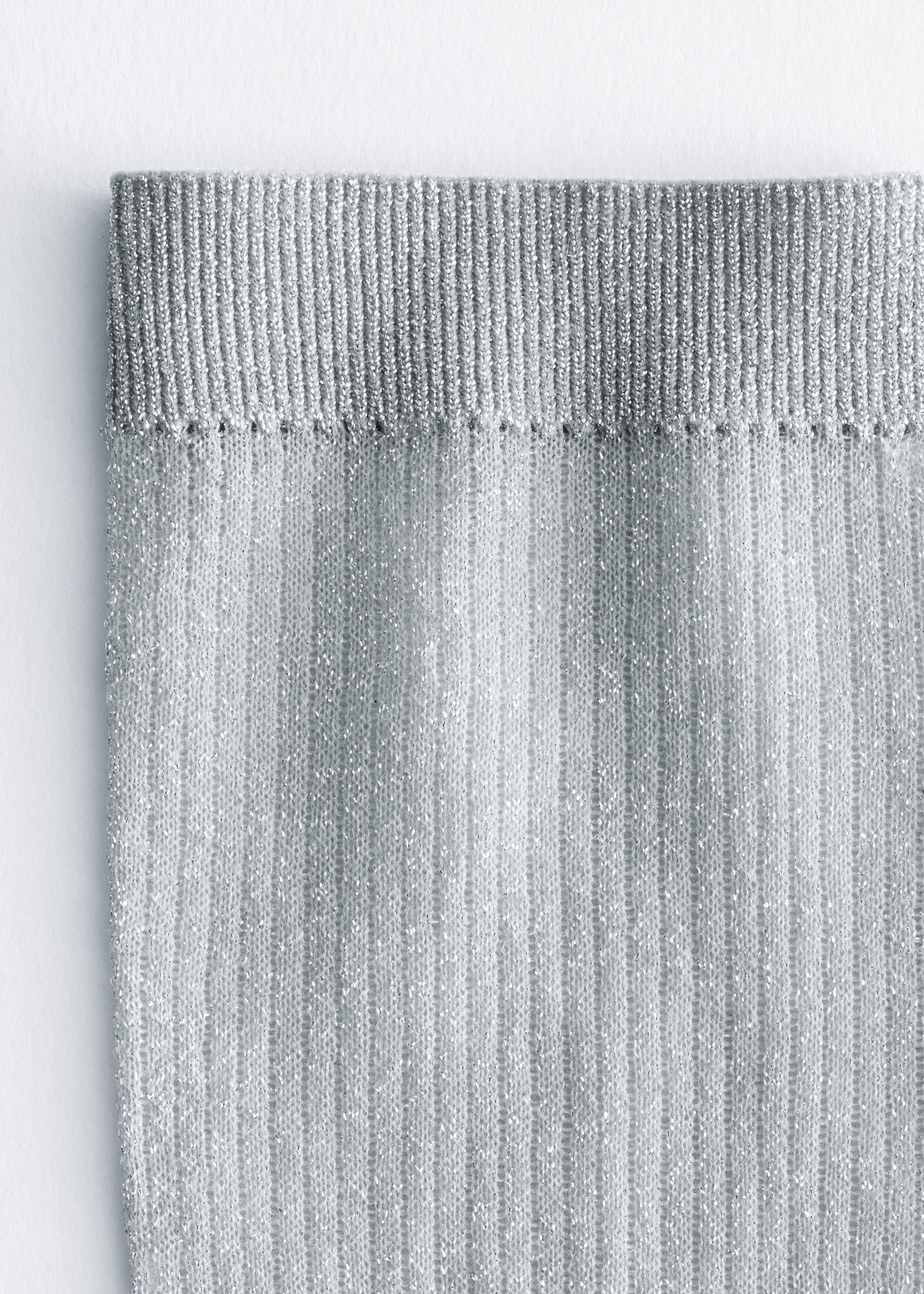A silver metallic sock showcases a finely ribbed top cuff and a vertically ribbed body woven with shimmering threads. The close-up view highlights the textured, glittery knit fabric of the sock.