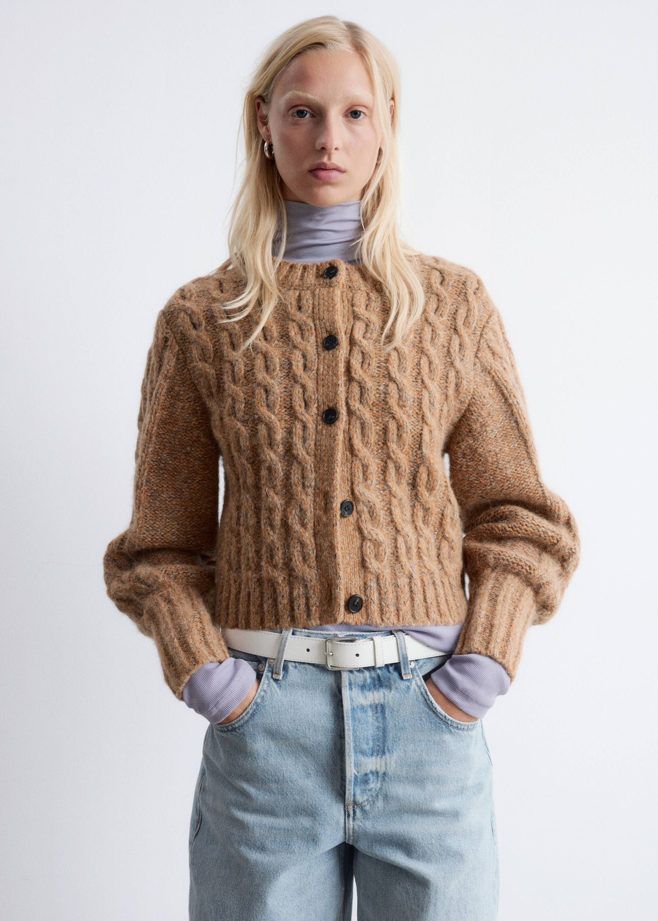 Agrandir l'image: Un cardigan à torsades épais et marron présente une silhouette courte, un col rond, des manches bouffantes volumineuses et des finitions côtelées. Des boutons sombres descendent le long du devant tandis que le cardigan est porté par le mannequin, qui le superpose à un haut à col roulé violet clair et l'associe à un jean délavé clair et une ceinture blanche.