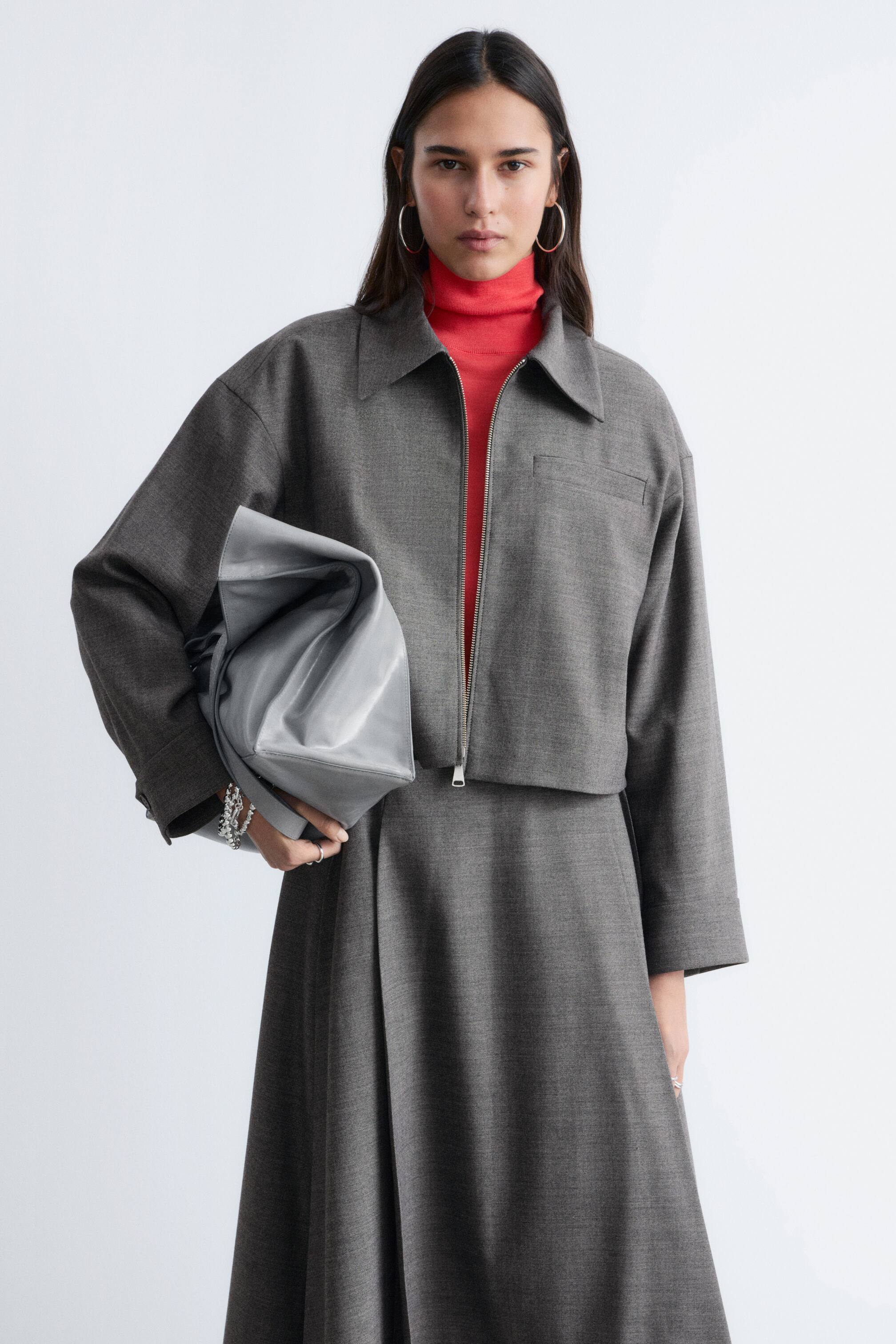 Grotere afbeelding bekijken: Een grijze bijpassende set, bestaande uit een kort jasje met een volledige ritssluiting en een puntige kraag, en een midi-rok, wordt gepresenteerd. Onder het jasje wordt een rode coltrui gedragen en een zachte, lichtgrijze tas wordt vastgehouden.