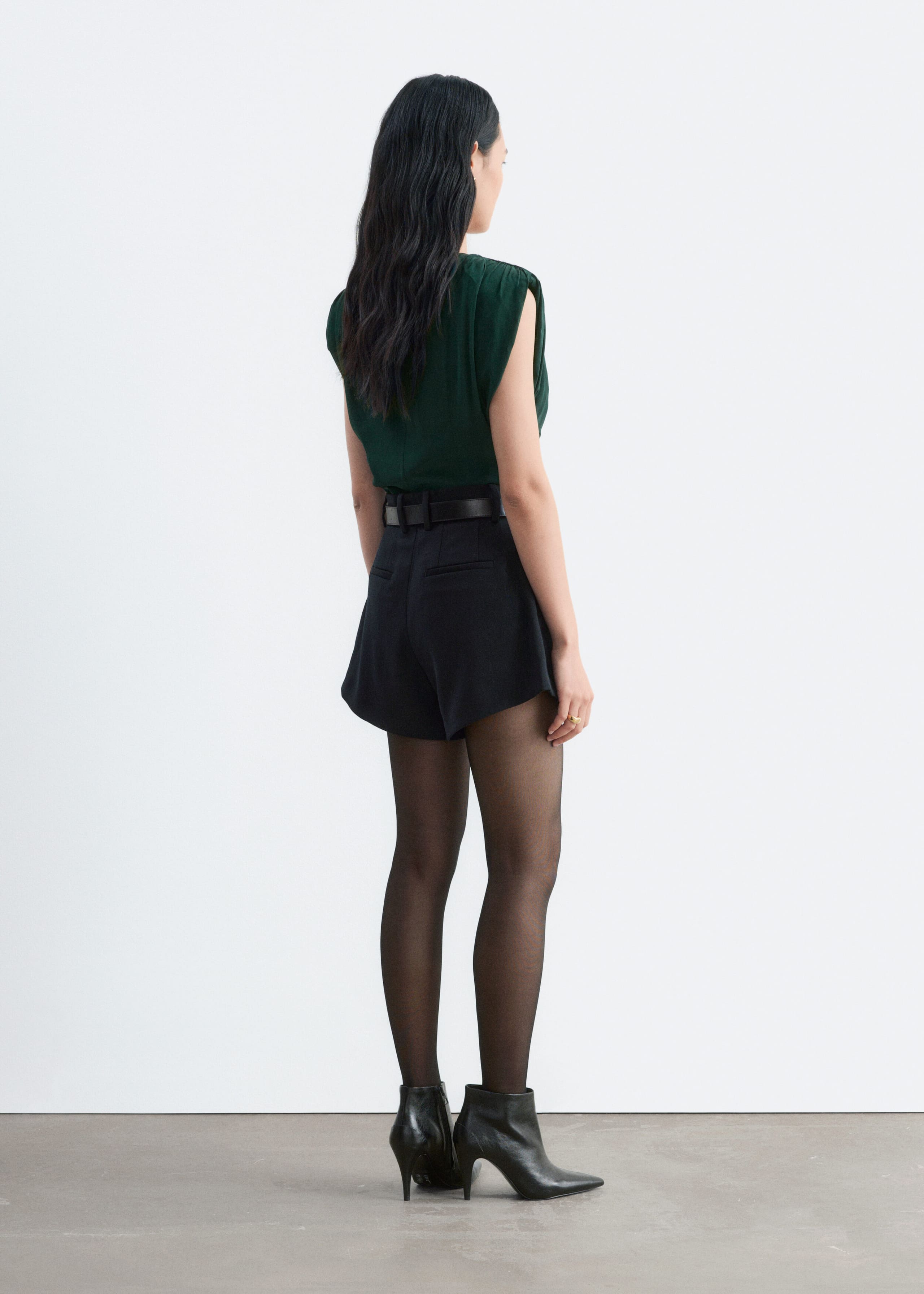 A dark green sleeveless top with ruched shoulders is worn by the model, tucked into high-waisted black shorts with a belt. The outfit is styled with sheer black tights and black heeled ankle boots, shown from a rear three-quarter angle.