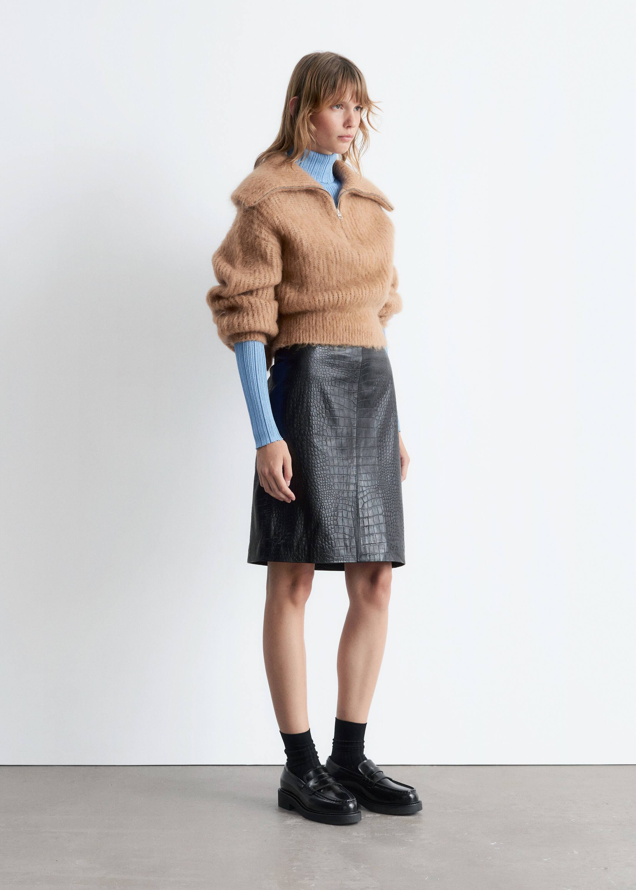 Grösseres Bild ansehen: Ein kamelfarbener, flauschiger Strick-Half-Zip-Pullover mit breitem Kragen und voluminösen Ärmeln ist über einem hellblauen gerippten Rollkragenpullover geschichtet, dessen lange Ärmel aus den Bündchen des Pullovers hervorragen. Ein schwarzer Minirock mit Krokodilprägung aus Kunstleder ergänzt den kurzen Pullover, während schwarze Loafer mit dicker Sohle und schwarze Socken das Ensemble vervollständigen.
