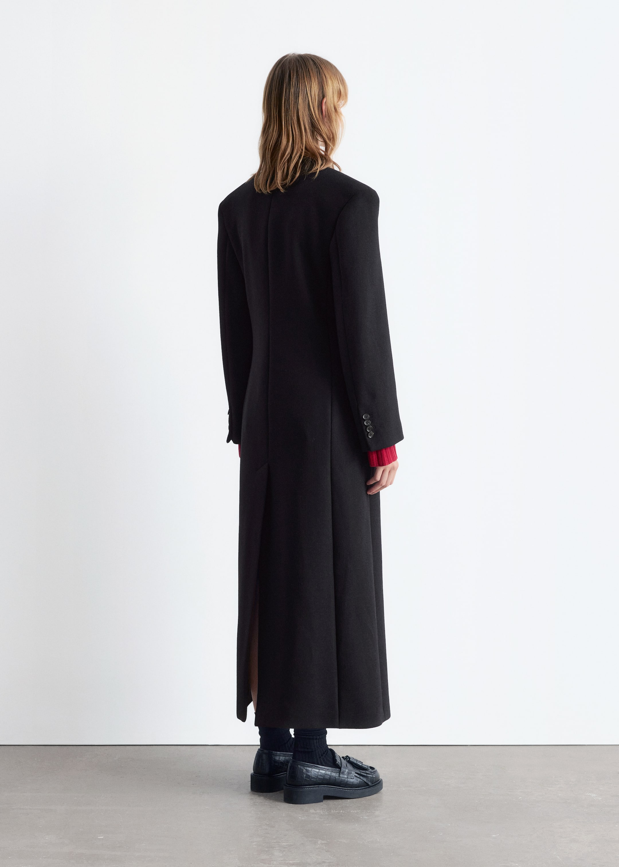View larger image: A black wool-blend coat with a long, straight silhouette, padded shoulders, buttoned cuffs, and a long back vent is being worn by the model, viewed from the back. A red long-sleeved garment is visible layering beneath its sleeves, and the model is standing, wearing dark loafers and socks.