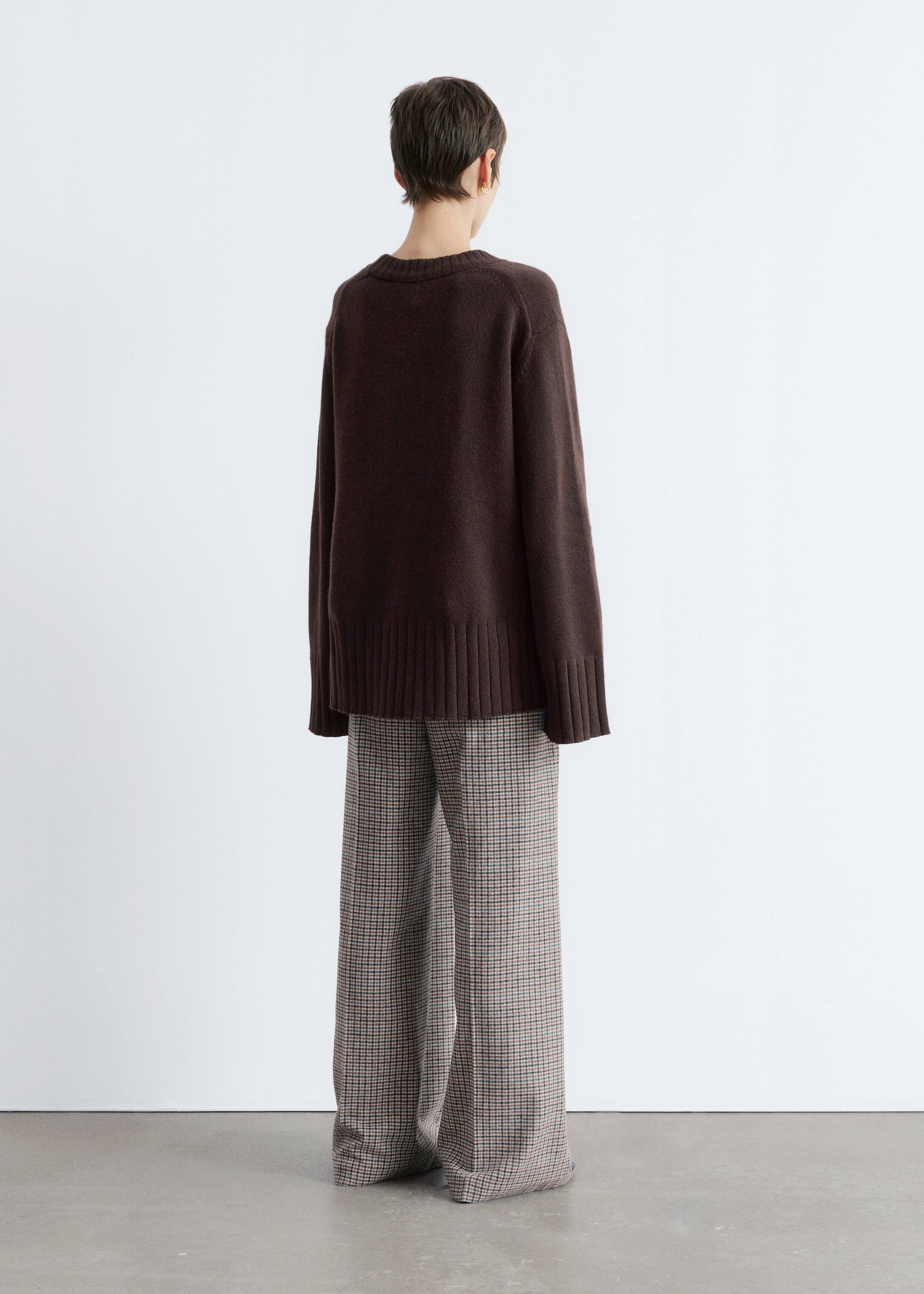 View larger image: A dark brown crewneck sweater is being presented from the back, showcasing its relaxed fit, ribbed hem, and ribbed cuffs. It is being styled over wide-leg trousers featuring a brown and beige plaid pattern.
