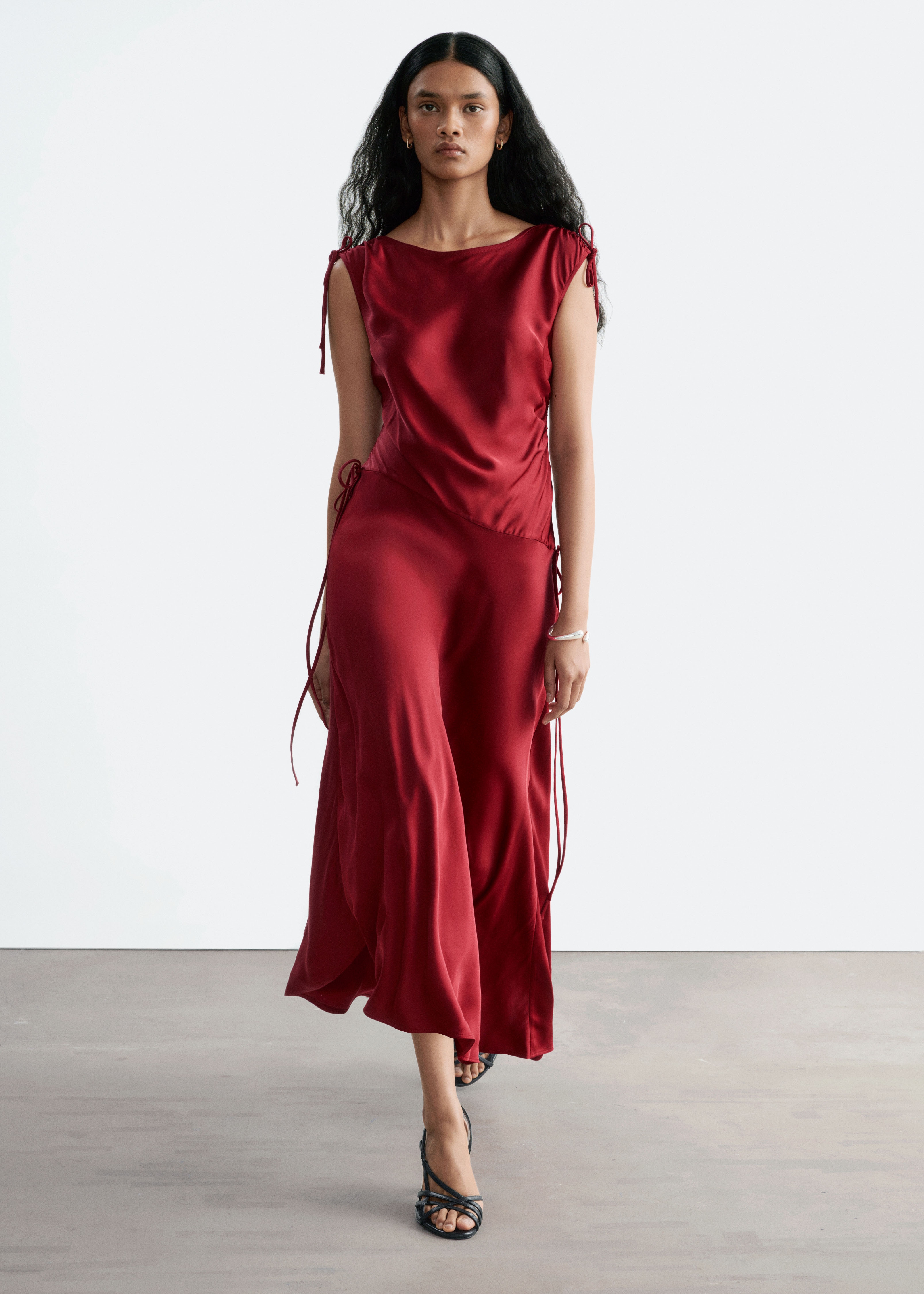 Um vestido midi de cetim vermelho está pendurado de frente, destacando um decote barco com alças ajustáveis com cordão e longos laços pendentes. O vestido apresenta uma cintura descaÃda assimétrica franzida que desce até a uma bainha drapeada assimétrica.