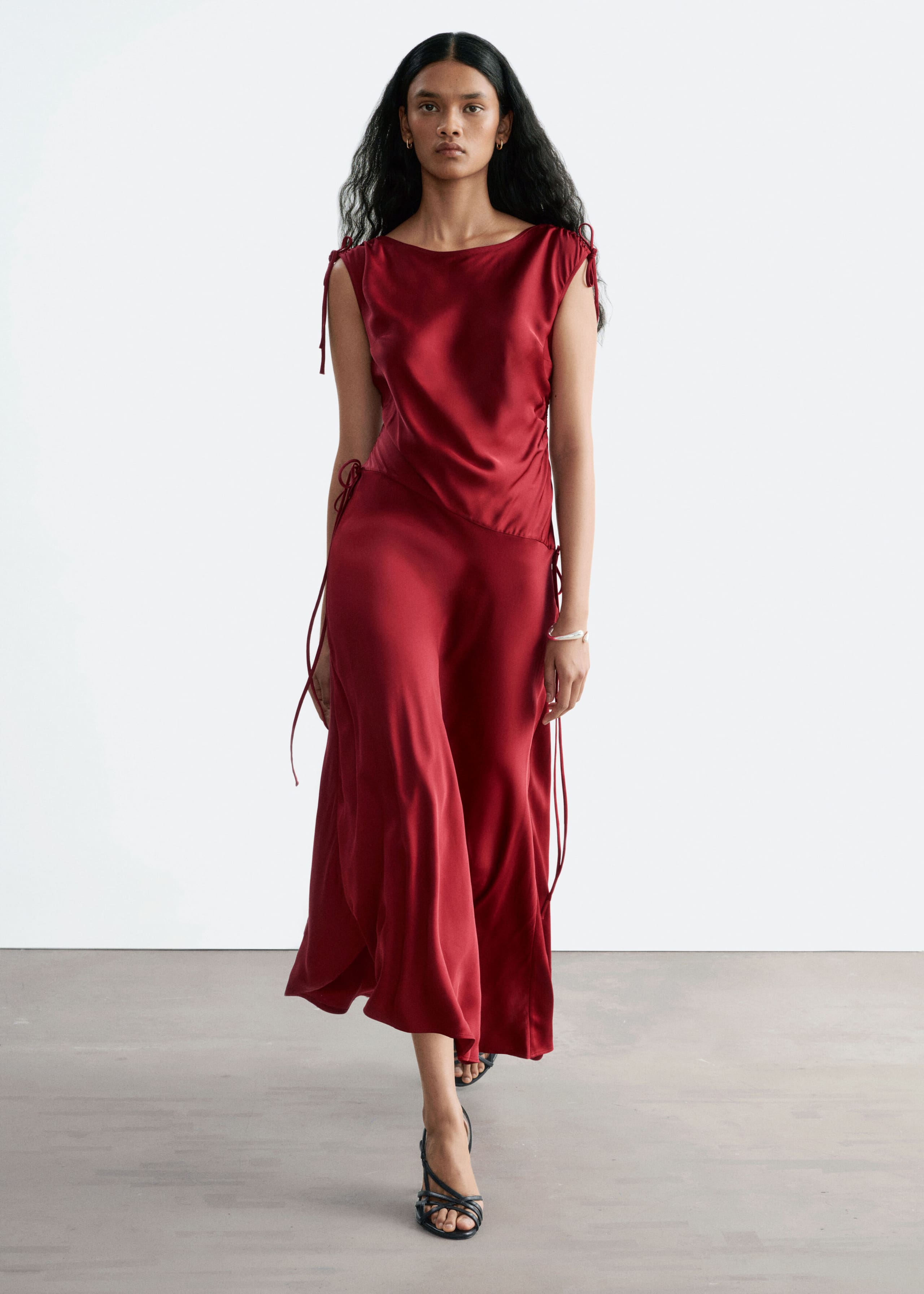 A red satin draped dress featuring drawstring ties at the shoulders and an asymmetric dropped waist is worn by the model. The dress has a fluid, asymmetric hemline and is styled with black strappy sandals.