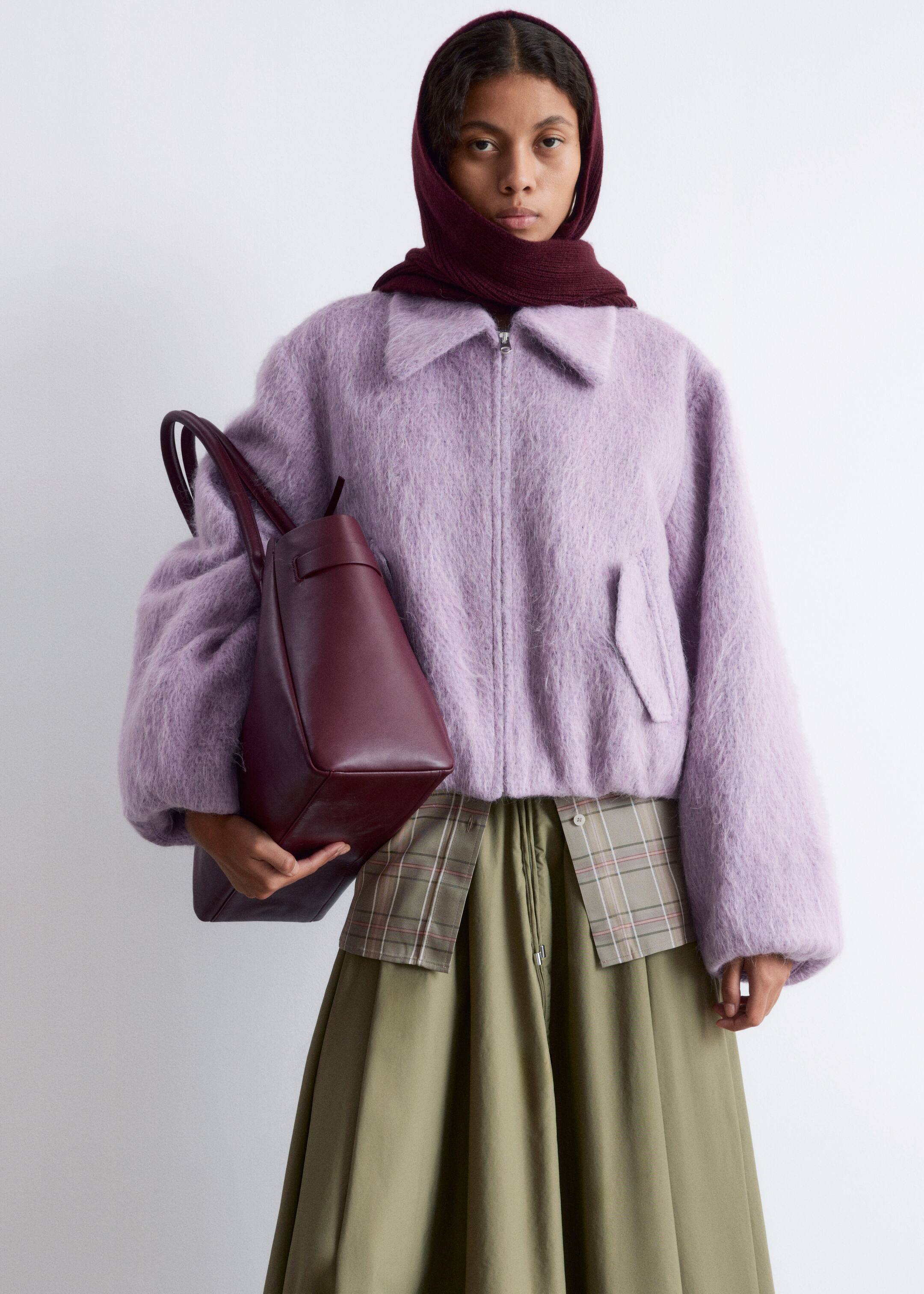 Visualizza immagine più grande: Una giacca bomber corta e soffice di colore viola chiaro presenta un colletto classico, spalle cadenti, maniche ampie, tasche anteriori con patta e chiusura a tutta zip, con il suo orlo elasticizzato che si posiziona sopra una gonna a pieghe. Una borsa tote in pelle strutturata color bordeaux con due manici superiori è tenuta nel gomito del braccio, posizionata contro il lato sinistro della giacca.