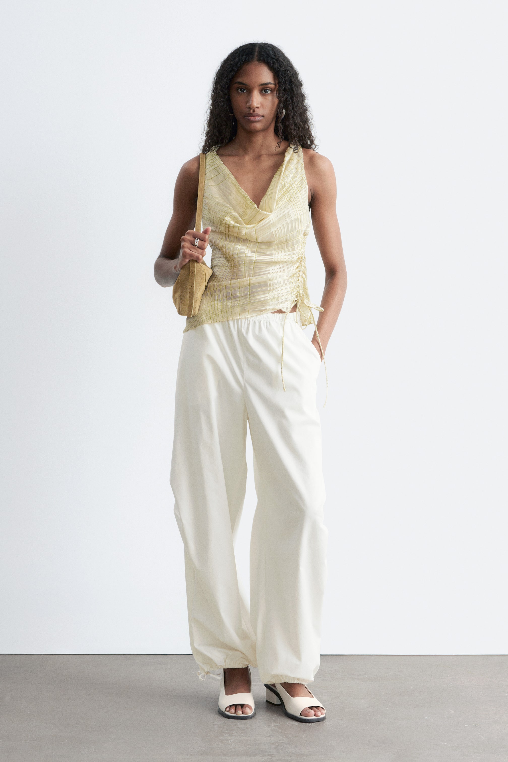 Ver imagem maior: Um top sem mangas de cor amarela clara, com padrão de rede transparente, gola drapeada e cordões laterais está a ser apresentado, combinado com calças largas de cor creme com tornozelos franzidos por cordões e sapatos de salto bloco abertos na frente de cor creme com solas pretas. Uma mala de ombro retangular de cor castanha está a ser segurada, todos os itens estão virados para a frente.