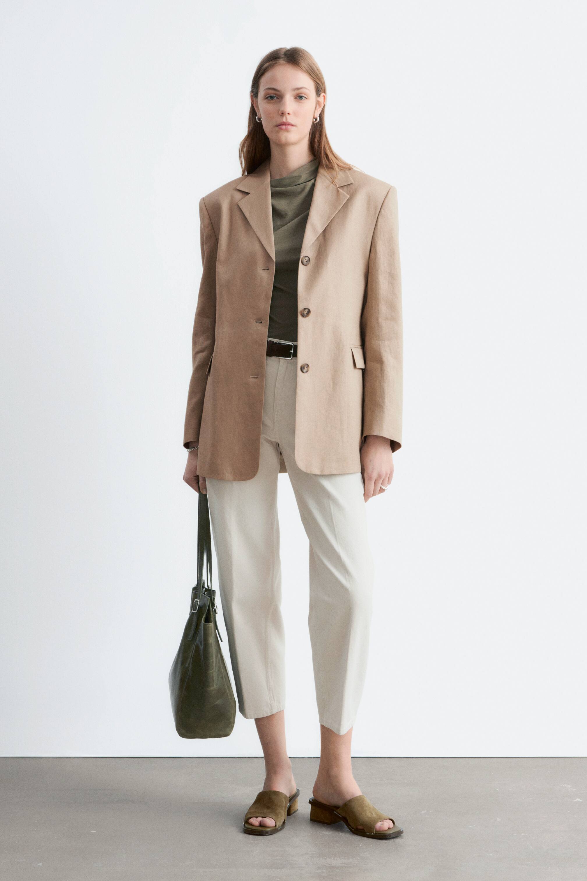 Se større billede: En beige blazer præsenterer en afslappet pasform med strukturerede skuldre, notch-revers og tre mørke knapper på frontlukningen. Den har to påsatte lommer og bæres over en mørk olivengrøn højhalset top.