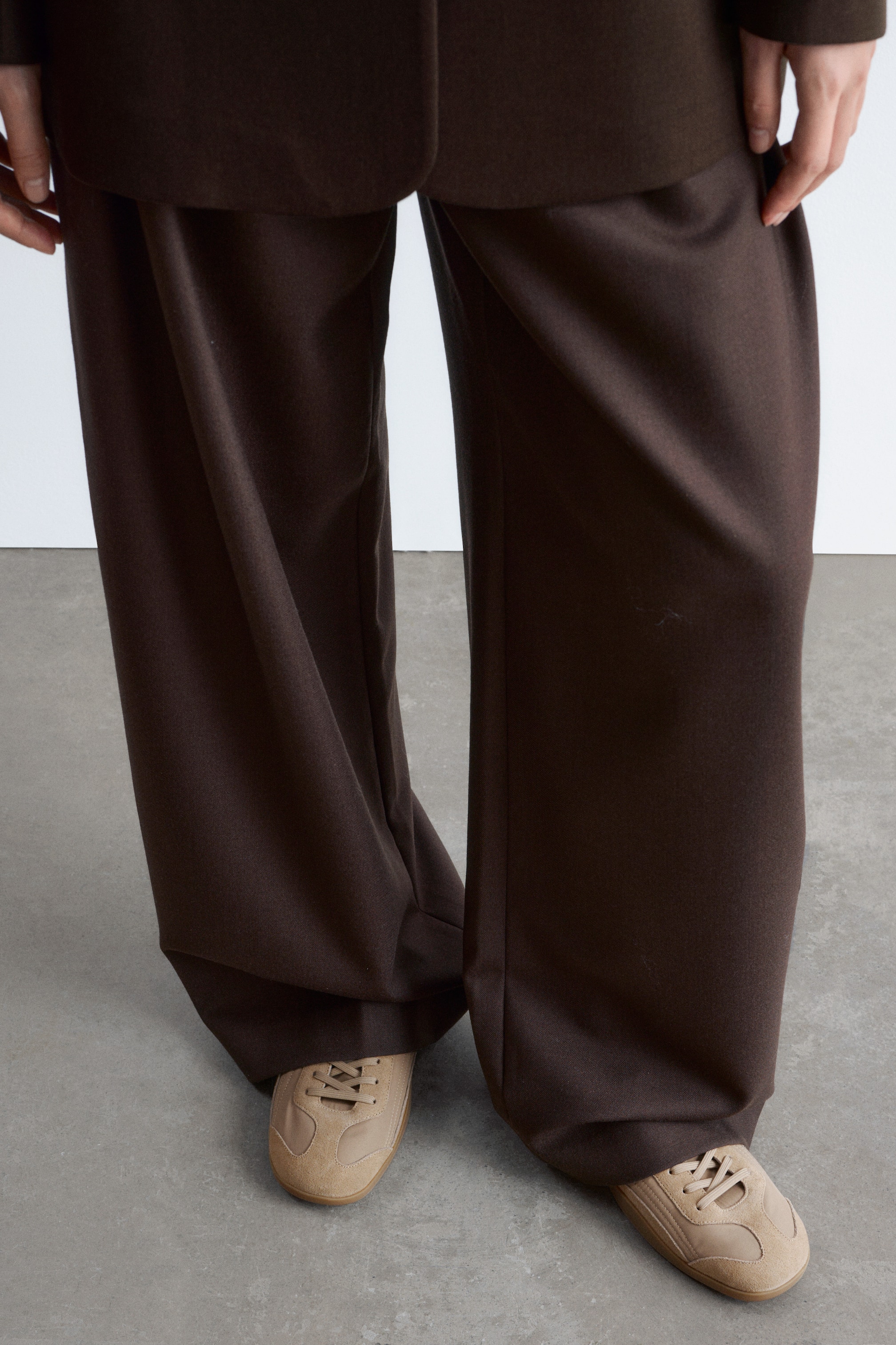 Grotere afbeelding bekijken: Beige leren vetersneakers, met een slank profiel, ballerina-geïnspireerde vorm, puntige amandelneus en golvende doorgestikte naden, worden gedragen door het model. De sneakers' patroonrubberen zool is zichtbaar, terwijl de wijde bruine broek van het model over de bovenkant van de sneakers valt.