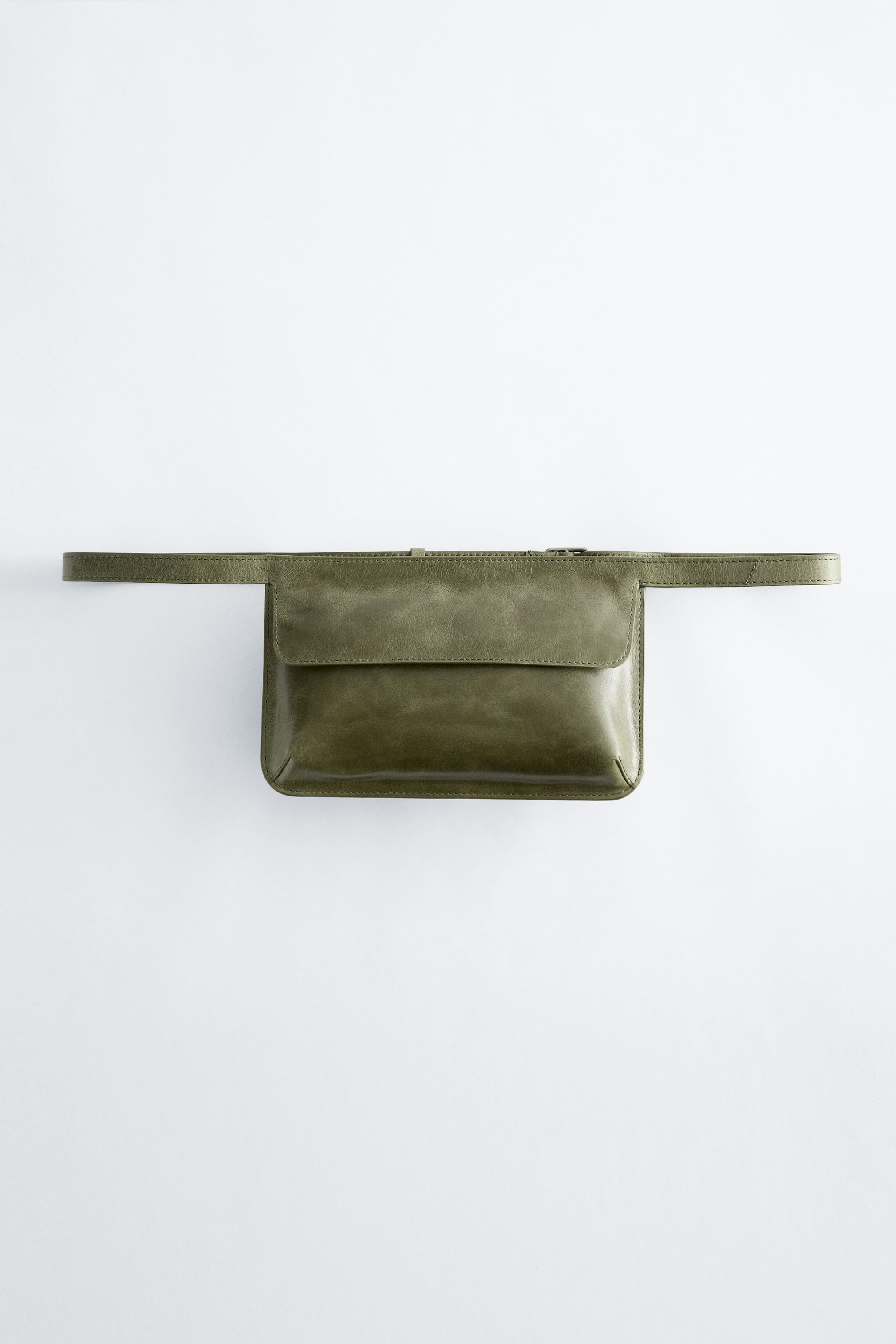 Se større billede: En olivengrøn læderbæltetaske viser sin front. Den har en rektangulær pung med en synlig frontklaplukning, en lynlås langs dens øverste kant og en tynd rem, der strækker sig vandret fra begge sider.