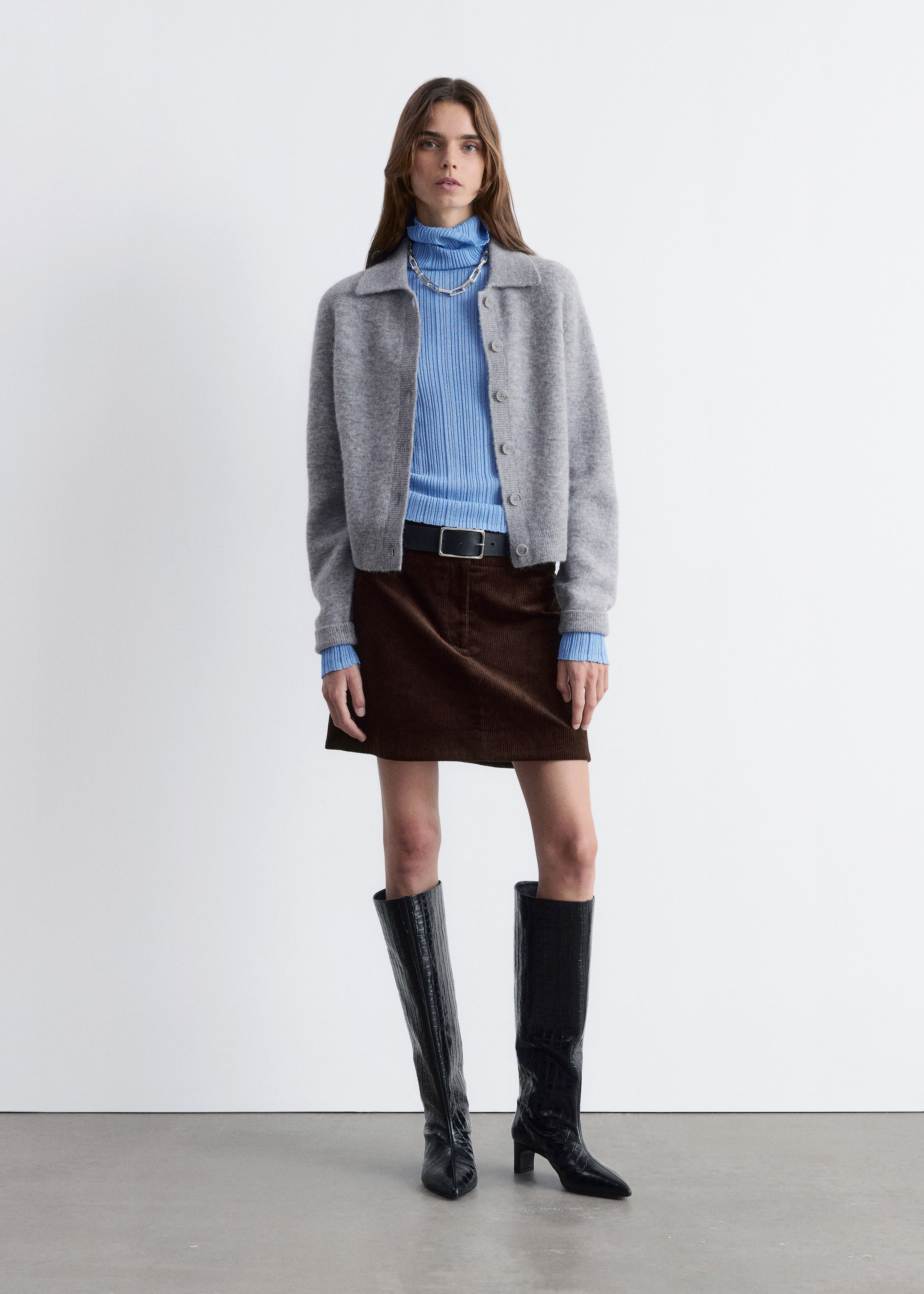 Agrandir l'image: Une tenue de face présente un cardigan tricoté gris clair avec un col classique et une patte de boutonnage, porté ouvert sur un pull à col roulé côtelé bleu clair. Une mini-jupe en velours côtelé marron foncé est portée avec une ceinture noire, stylisée avec des bottes noires à hauteur de genou avec un bout pointu et un talon bloc, et un collier chaîne en argent massif est porté sur le col roulé.