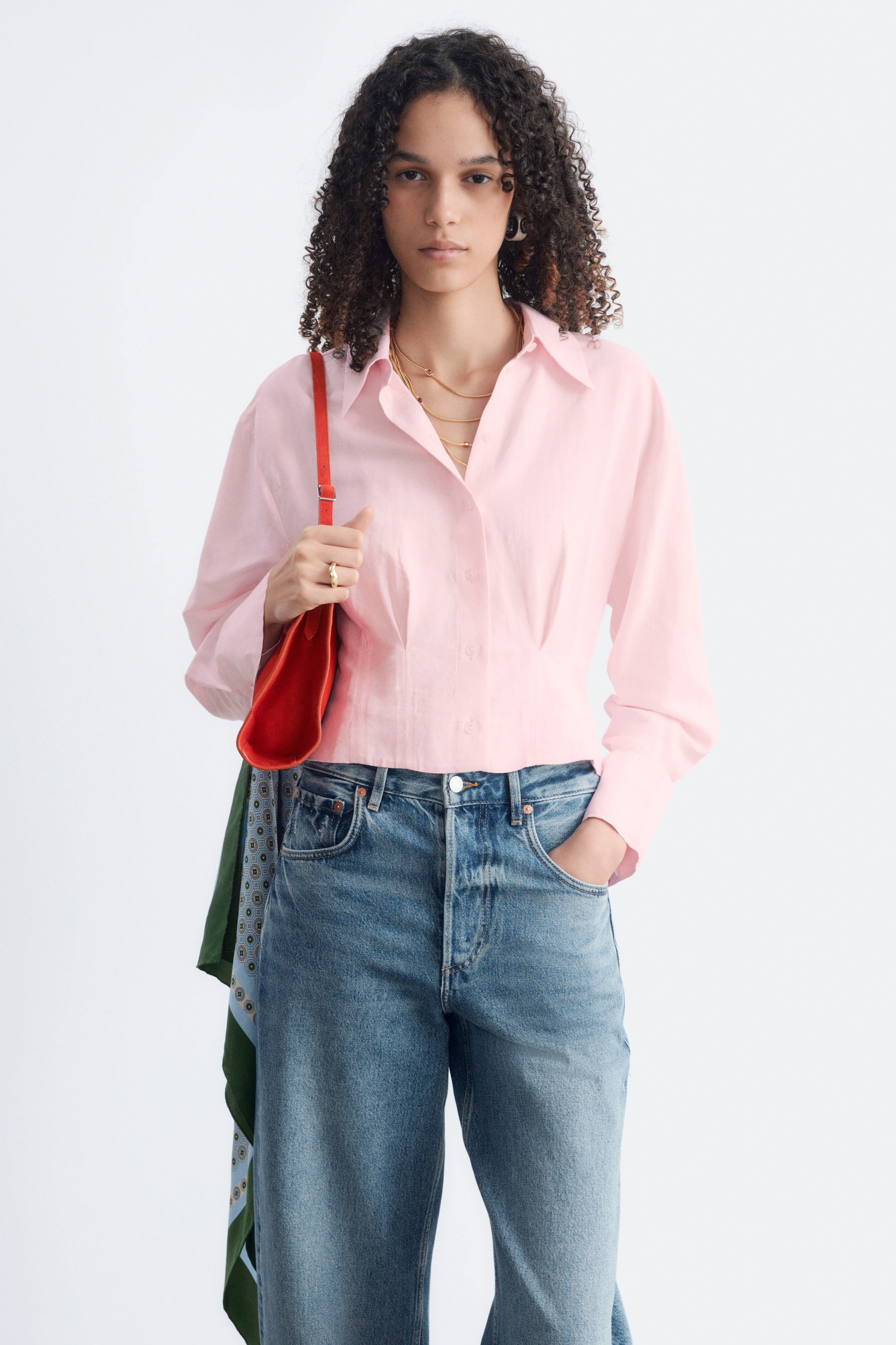 Agrandir l'image: Une chemise rose à manches longues présente un col classique rabattu, une patte de boutonnage sur le devant et une taille cintrée avec des plis détaillés. Le modèle porte la chemise, qui a une longueur raccourcie et un col ouvert.